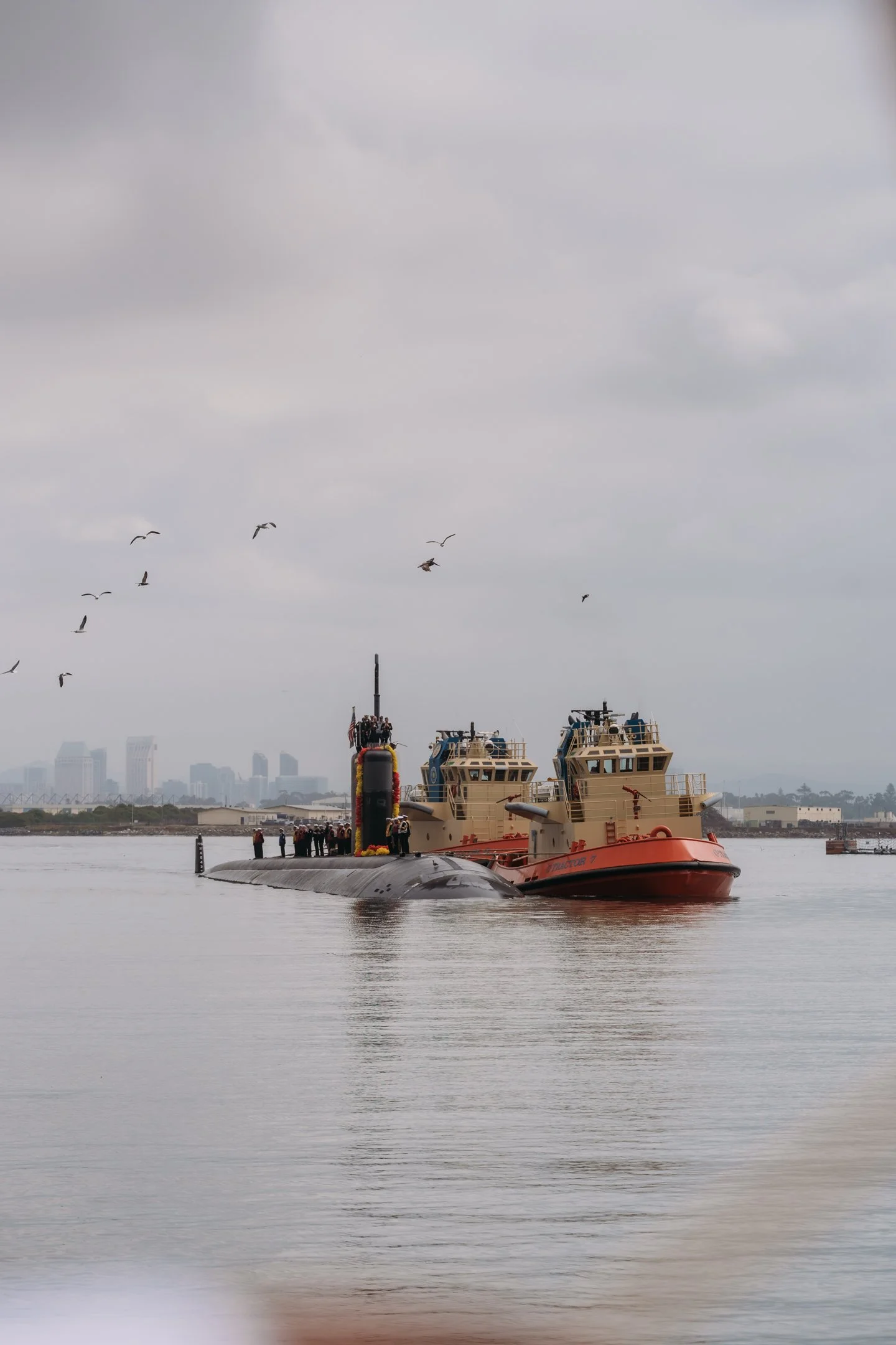 A submarine submerged in water with two tugboats on either side, and people standing on the submarine's deck, with a city skyline and seagulls flying overhead in the background.   San Diego Military Homecoming Photograph