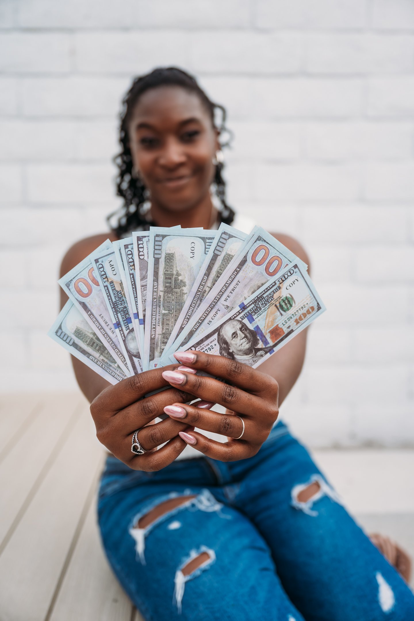 A woman with dark skin and curly hair sitting on a wooden floor holding fan of $100 bills towards the camera, with a blurred white brick wall in background. Alisha Mowry Photography Military, Brand, and Portrait Photographer San Diego CA