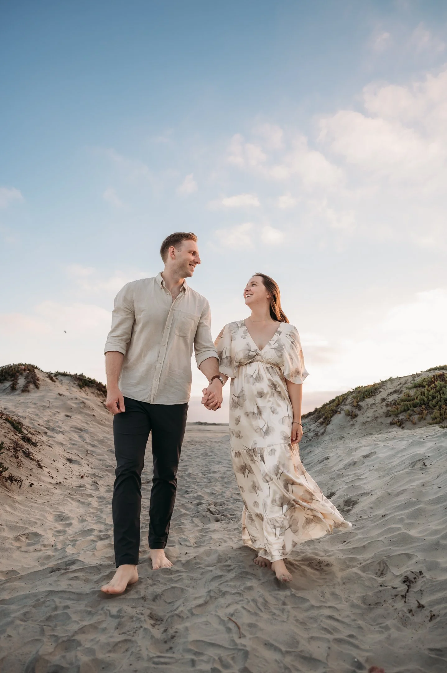 A happily smiling couple holding hands and walking barefoot on a sandy beach during sunset or sunrise.   Alisha Mowry Photography Military, Brand, and Portrait Photographer San Diego CA