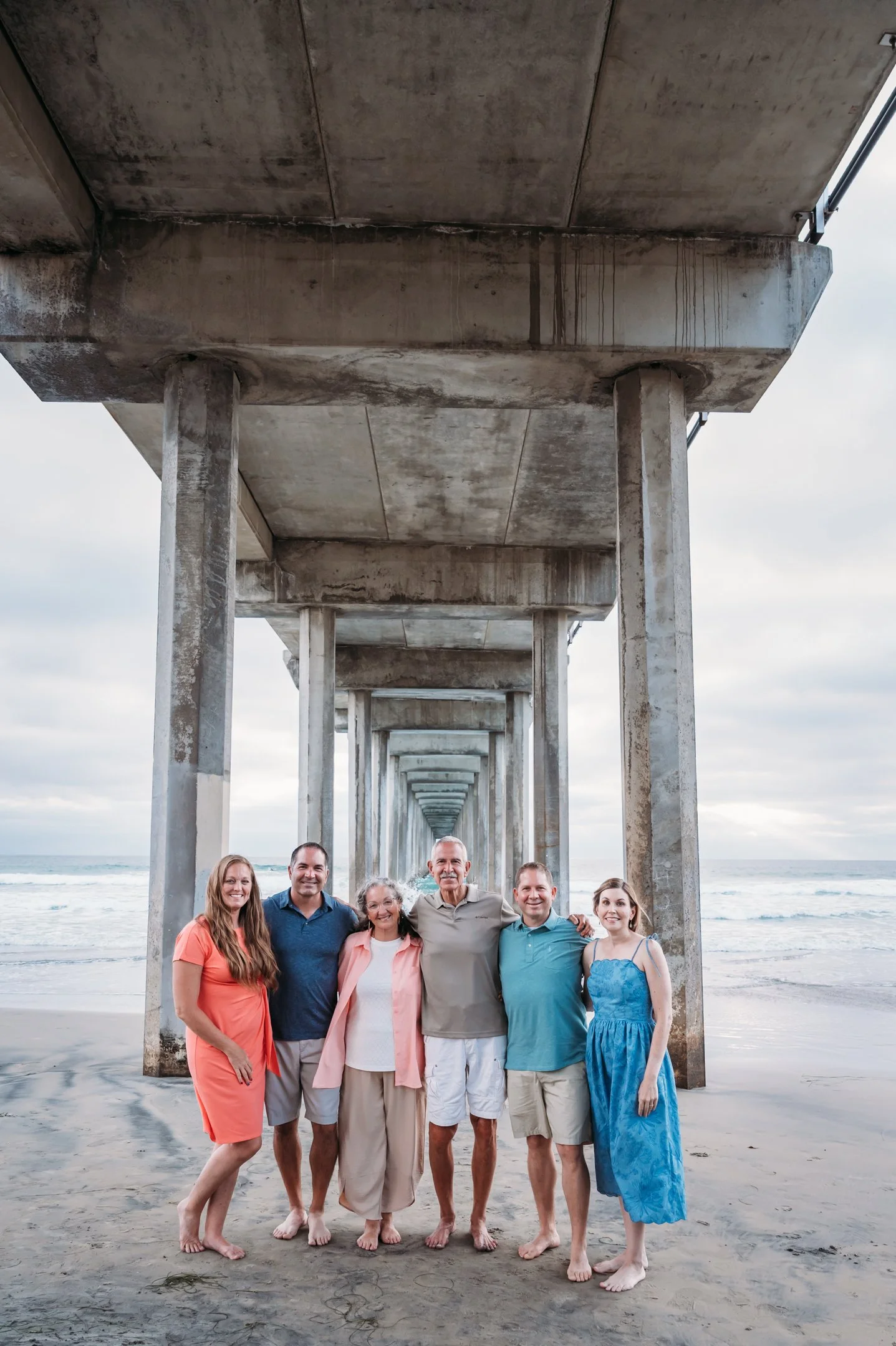 Group of six people standing on a sandy beach under a large concrete pier overcast sky