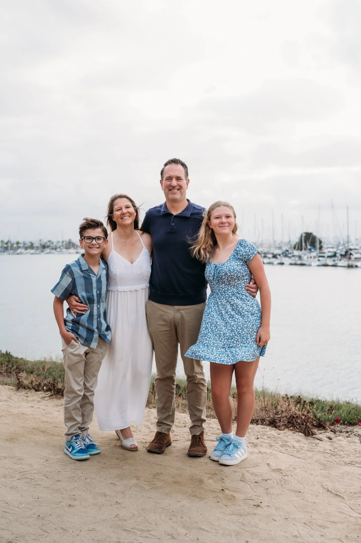 Family of four standing outdoors near a body of water with boats in the background, smiling at the camera. Alisha Mowry Photography Military, Brand, and Portrait Photographer San Diego CA