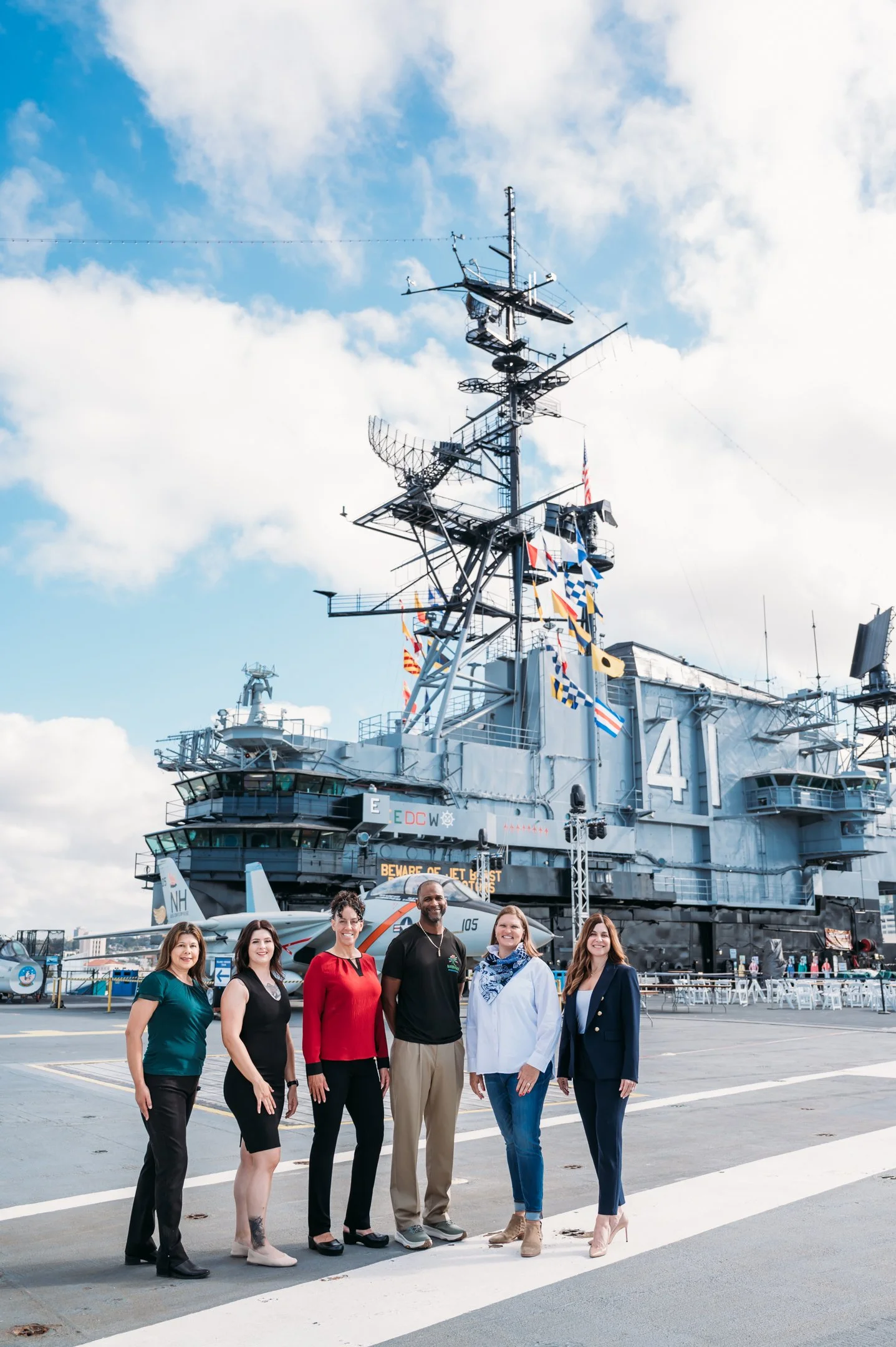 Group of six business professionals standing in front of the USS Yorktown aircraft carrier at a naval shipyard, with a cloudy sky above. Alisha Mowry Photography Military, Brand, and Portrait Photographer San Diego CA