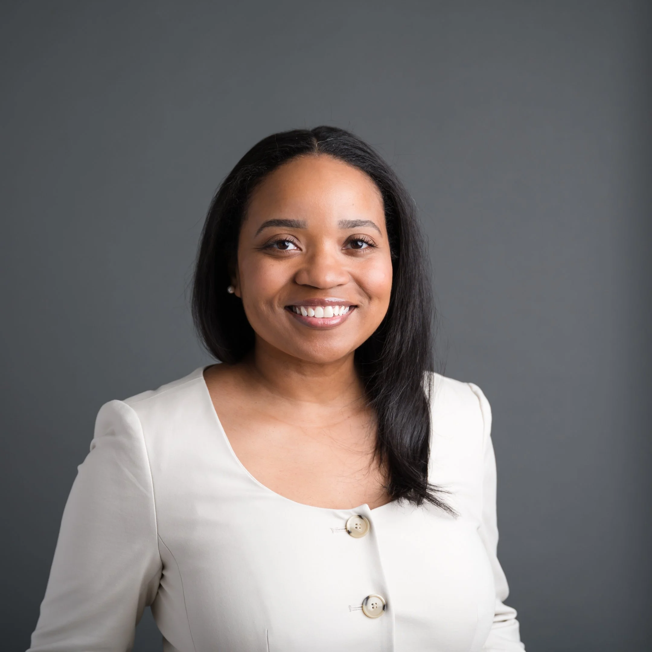 A professional woman with shoulder-length black hair, wearing a white blazer with two buttons, smiling against a gray background. Alisha Mowry Photography Military, Brand, and Portrait Photographer San Diego CA