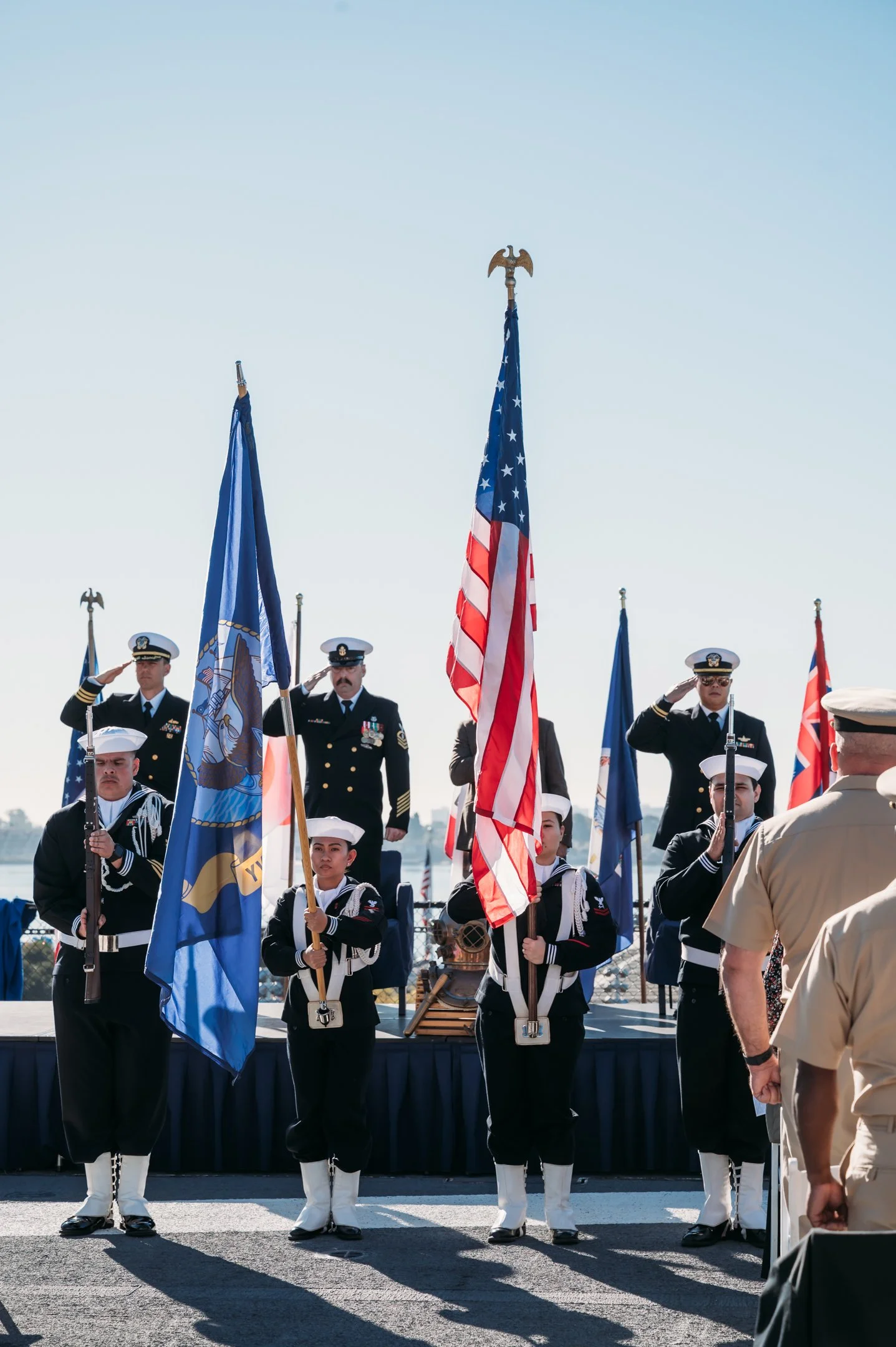Military personnel standing at attention during a flag ceremony, with service members holding American and other flags, and a decorated officer saluting in the background. uss midway military retirement ceremony 