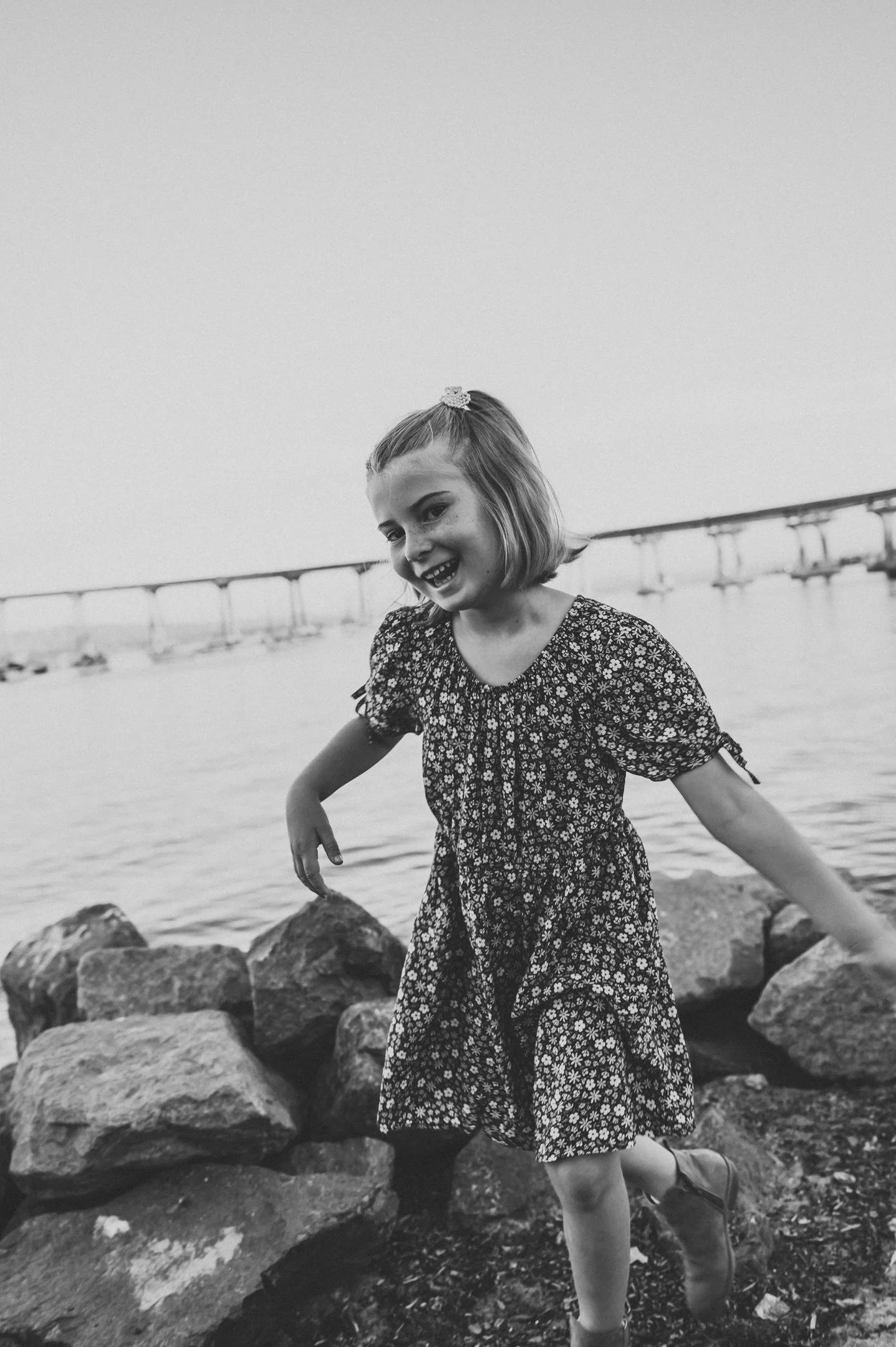 A young girl in a floral dress playing near rocks by the water, with a bridge in the background, seen in black and white. Alisha Mowry Photography Military, Brand, and Portrait Photographer San Diego CA