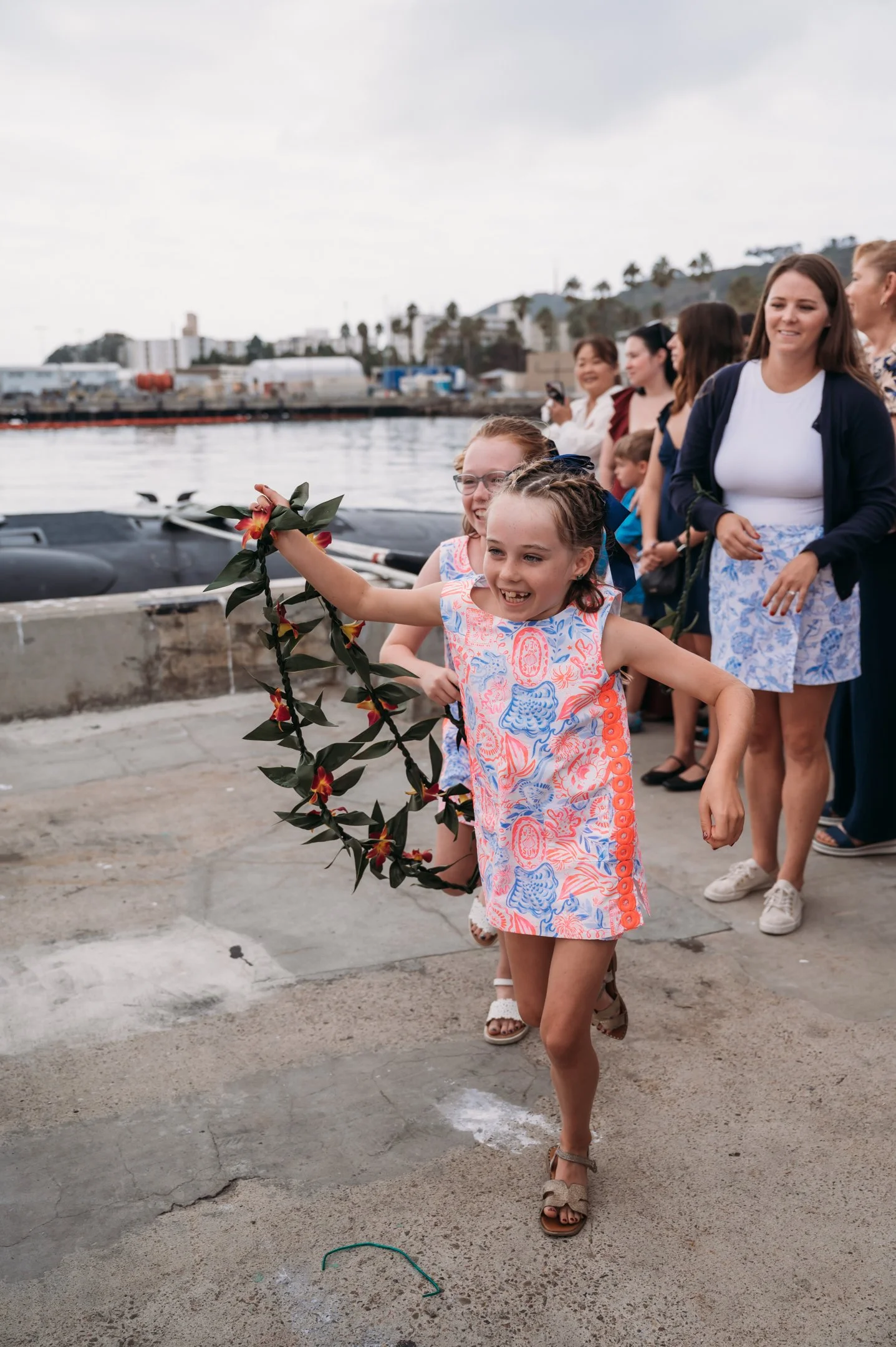 A young girl in a colorful dress holding a flower crown, running happily near a waterfront with a group of women and children behind her, likely during a celebration or festival. Alisha Mowry Photography Military Photographer San Diego