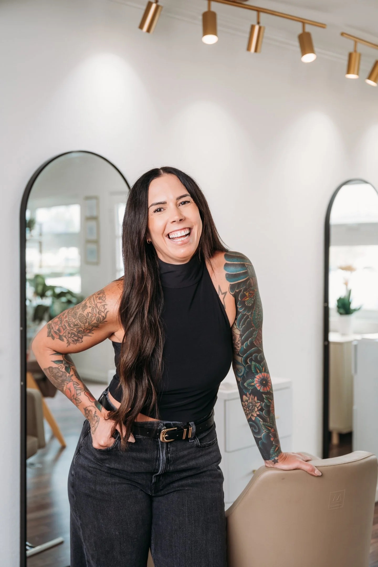 A woman with long dark hair and tattoos on her arms, wearing a black sleeveless top and black jeans, smiling and leaning on a chair in a modern, well-lit room with mirrors and decorative lighting. Alisha Mowry Photography Military, Brand, and Portrai