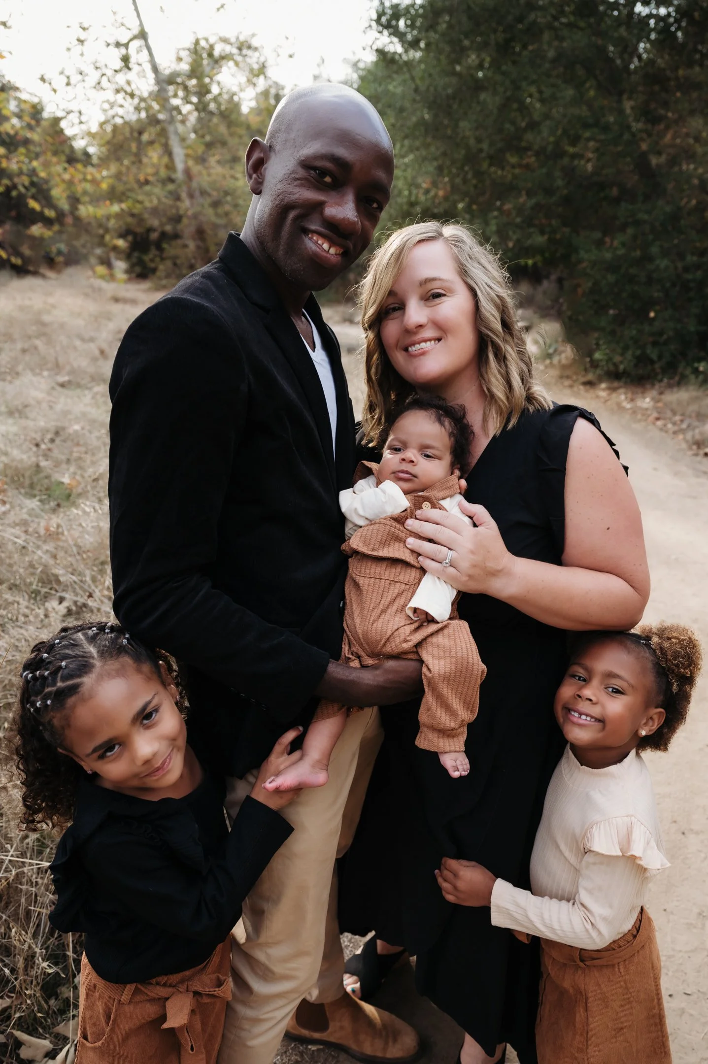A happy multiethnic family outdoors, with two adult women, a man, and three young girls, smiling and posing for a photo in a natural setting with trees in the background. Alisha Mowry Photography Military, Brand, and Portrait Photographer San Diego C
