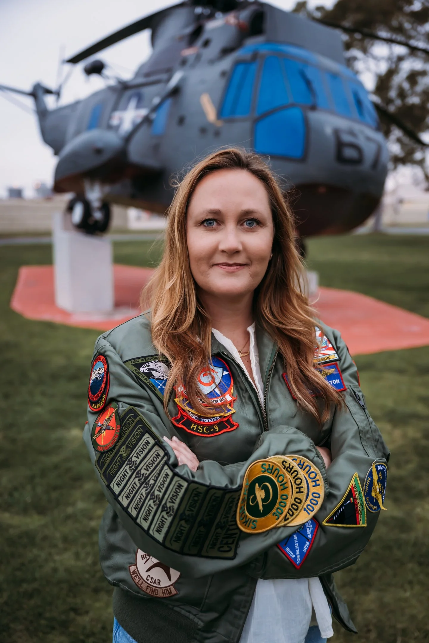 Woman in military jacket standing outdoors with arms crossed, in front of helicopter on display.