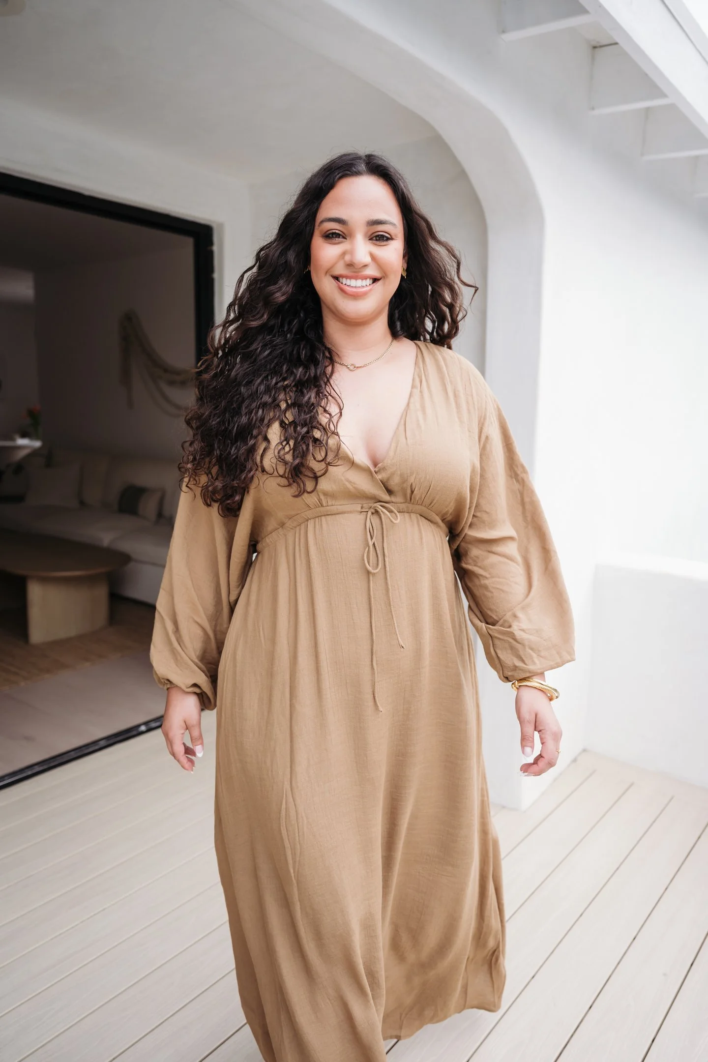 Smiling woman with long curly dark hair wearing a tan dress with a drawstring waist, standing on a light wooden porch with a white wall and stairs in the background. Alisha Mowry Photography Military, Brand, and Portrait Photographer San Diego CA