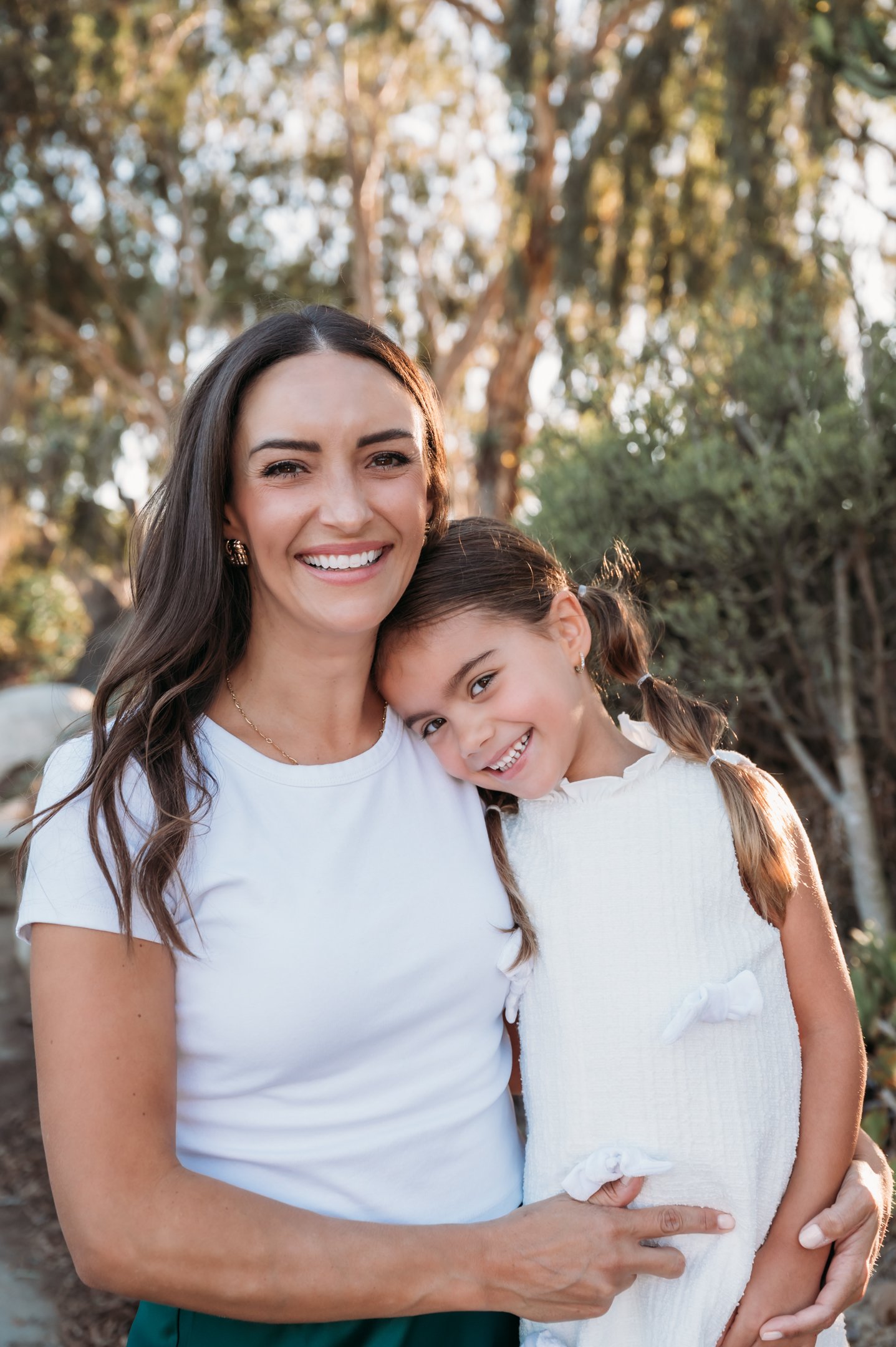 A smiling woman holding a young girl with pigtails, both in white tops, standing outdoors with trees in the background. Alisha Mowry Photography Military, Brand, and Portrait Photographer San Diego CA