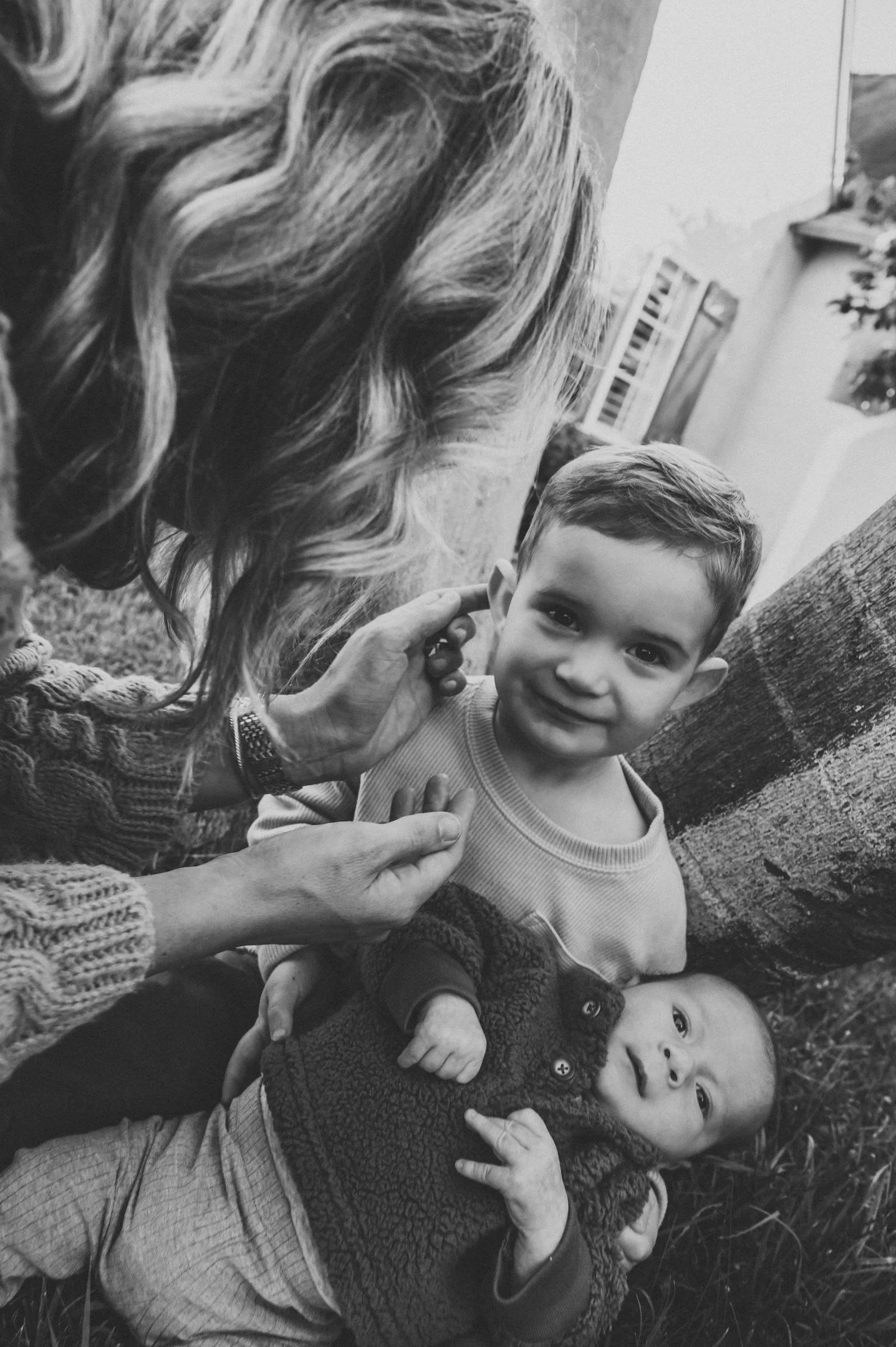 A black and white photo of a woman with curly hair gently touching a young boy's ear, who is smiling at the camera. The boy is leaning against a tree and holding a baby who is lying on the grass, looking up with a curious expression. Alisha Mowry Pho