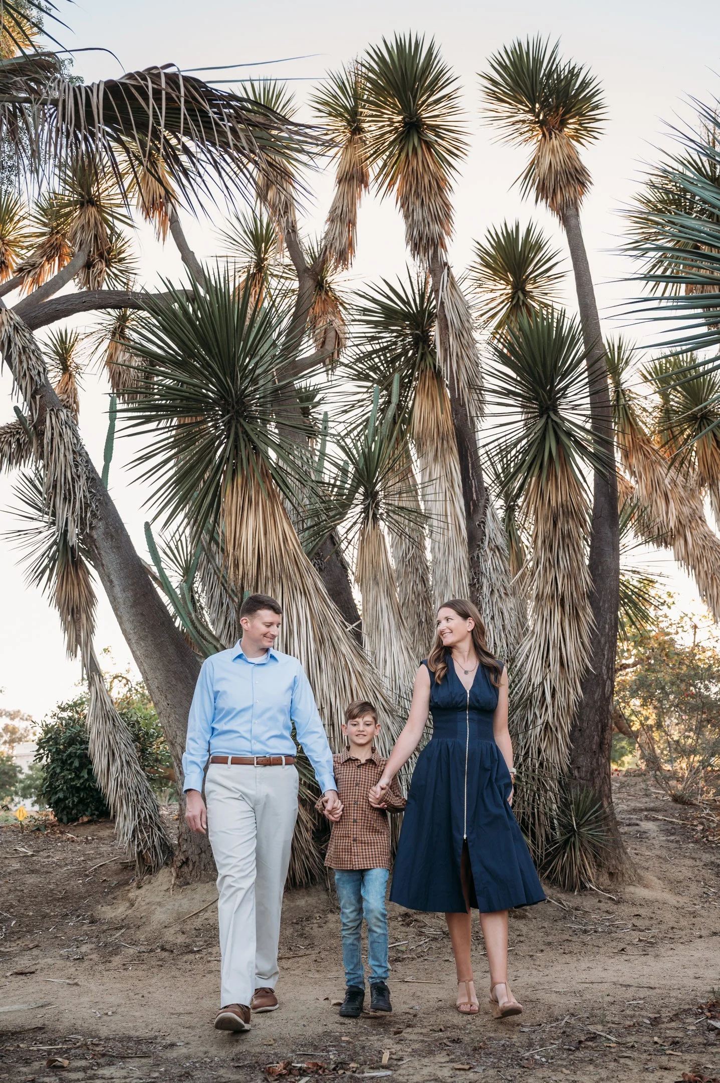 A family of three, a man, a woman, and a boy, holding hands and walking outdoors in front of large desert plants with long, spiky leaves at sunset. Alisha Mowry Photography Military, Brand, and Portrait Photographer San Diego CA