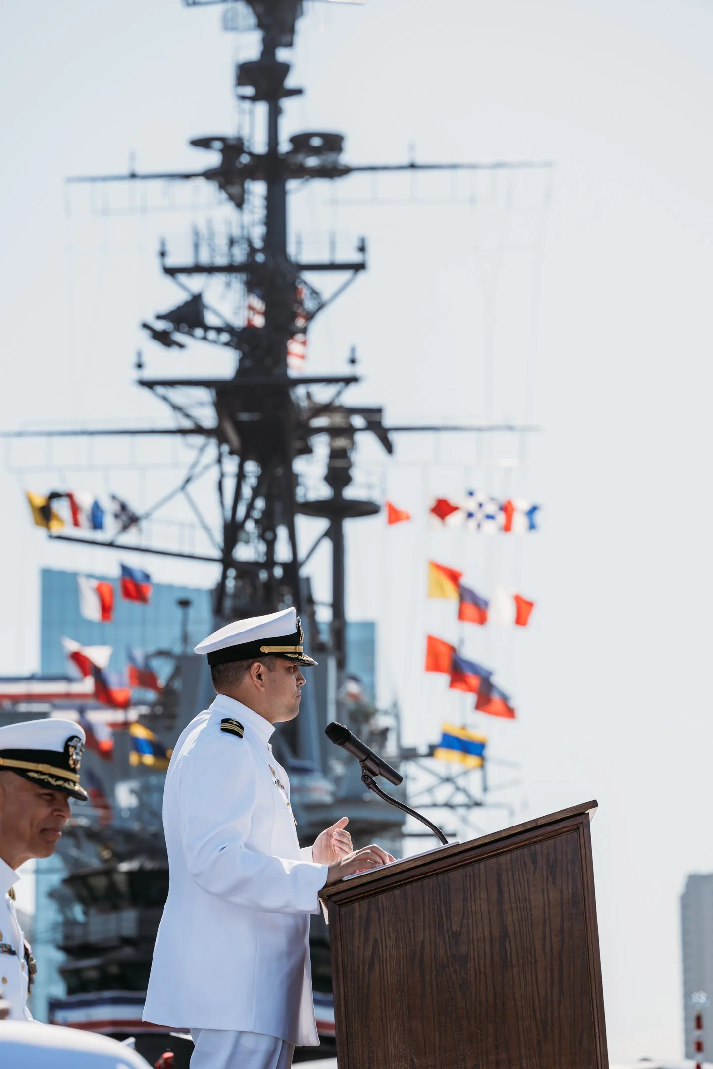 A naval officer in white uniform speaking at a podium during a ceremony, with a battleship and signal flags in the background. Alisha Mowry Photography Military Photographer San Diego