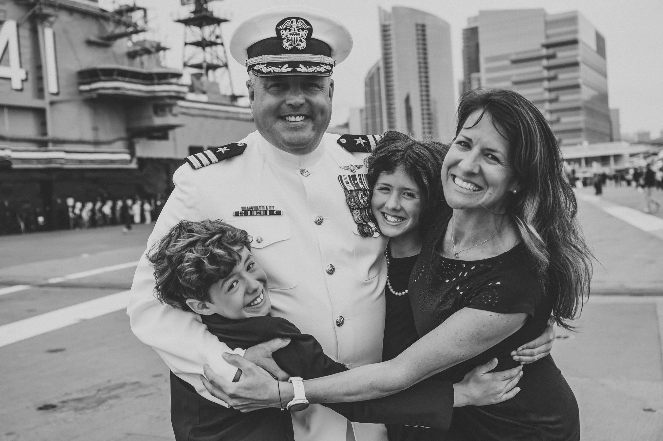 Black and white photo of a family hugging before a military officer in uniform with medals, on an urban street with tall buildings in the background. Alisha Mowry Photography Military Photographer San Diego