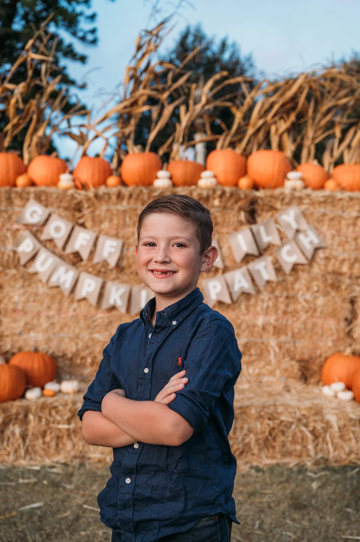 A young boy stands with arms crossed, smiling in front of a fall-themed background with pumpkins, gourds, and hay bales, with a 'Goofy PumPkin Patch' banner. Alisha Mowry Photography Military, Brand, and Portrait Photographer San Diego CA