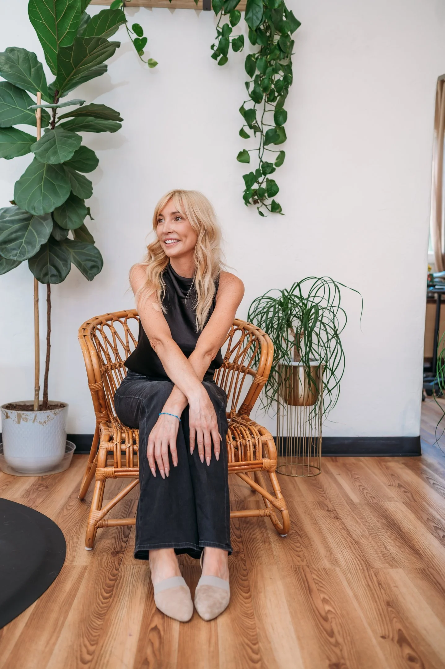 A woman with blonde hair sitting on a wicker chair in a room decorated with green houseplants and wooden flooring. Alisha Mowry Photography Military, Brand, and Portrait Photographer San Diego CA