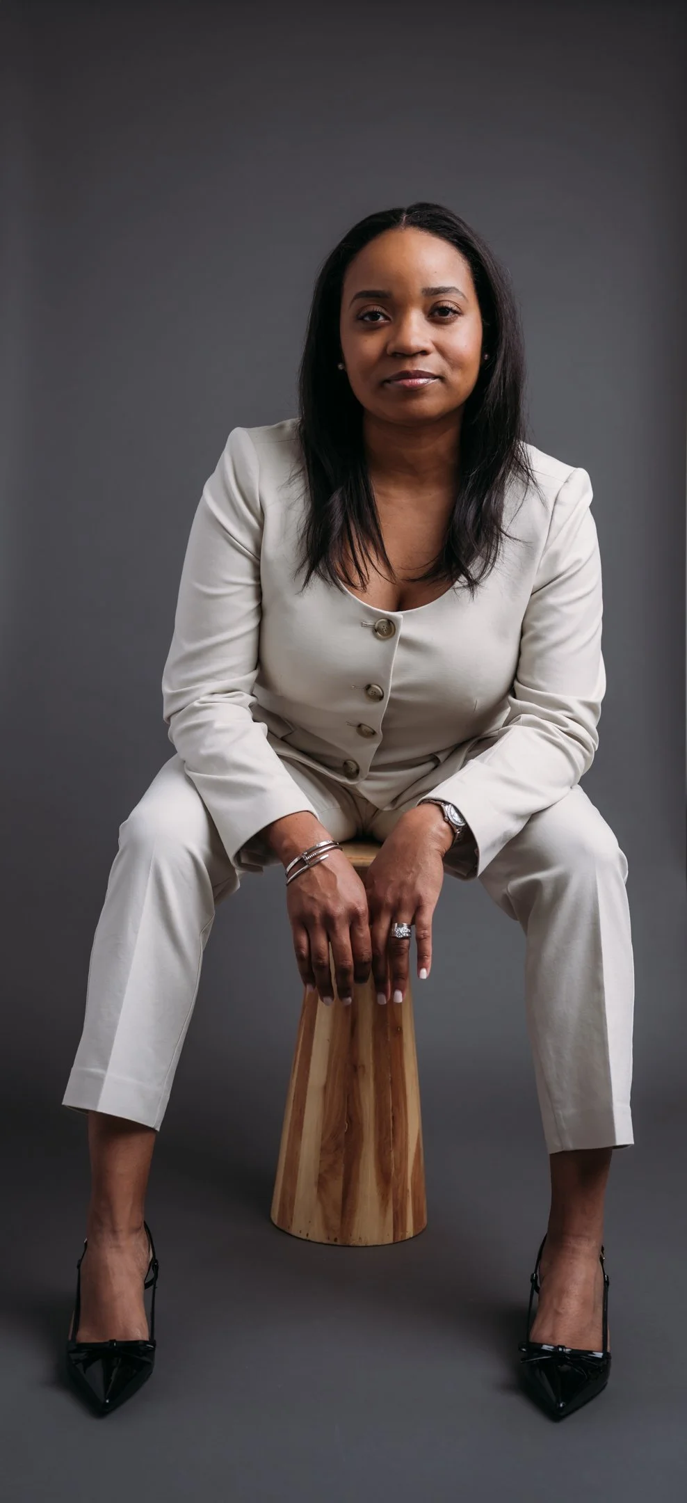 A woman in a white suit sitting on a wooden stool against a dark gray background. Alisha Mowry Photography Military, Brand, and Portrait Photographer San Diego CA