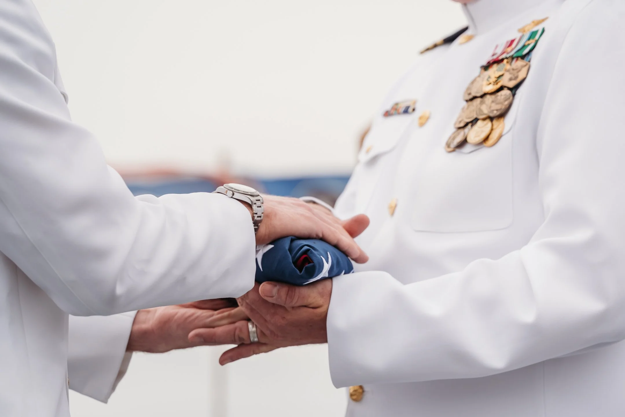 A military officer in white uniform with medals receiving a folded American flag from another person in a white coat, with hands overlapping on the flag. retirement ceremony on the use midway 