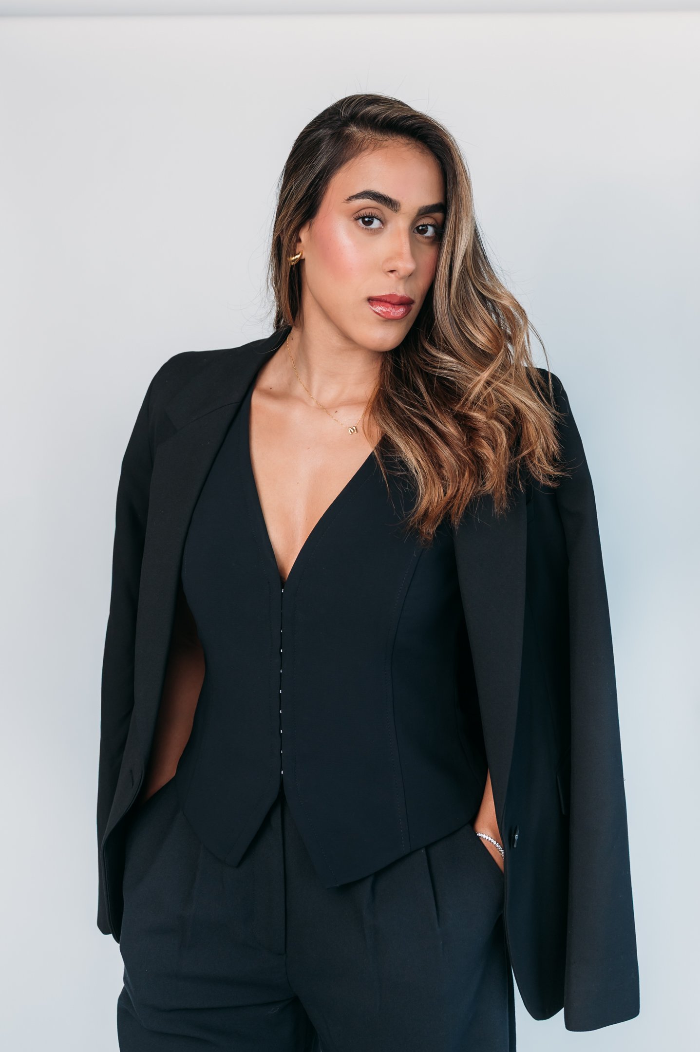A woman with long, wavy brown hair wearing a black blazer draped over her shoulders and a black V-neck blouse, standing against a plain white background. Alisha Mowry Photography Military, Brand, and Portrait Photographer San Diego CA