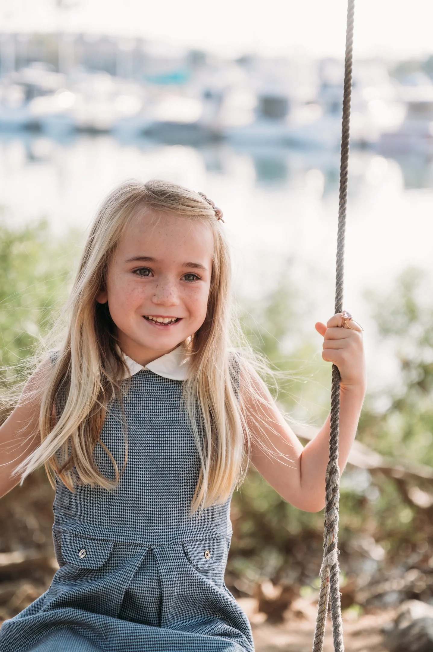 A young girl with long blonde hair, wearing a sleeveless dress with a white collar, is smiling and sitting on a swing near a body of water with boats in the background. Alisha Mowry Photography Military, Brand, and Portrait Photographer San Diego CA