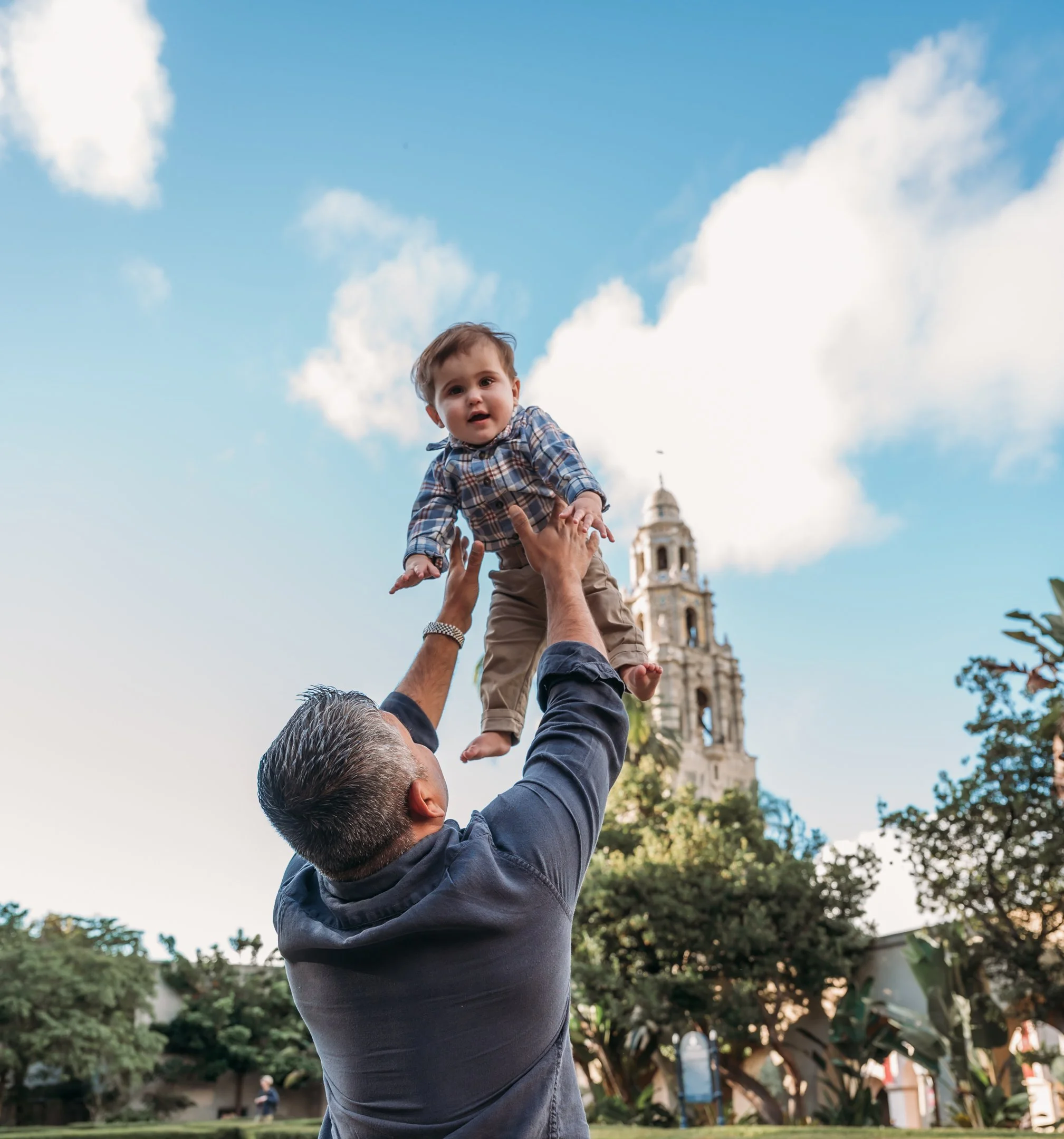 A man lifts a young boy into the air outdoors with a historic clock tower and trees in the background, blue sky with a few clouds. Alisha Mowry Photography Military, Brand, and Portrait Photographer San Diego CA