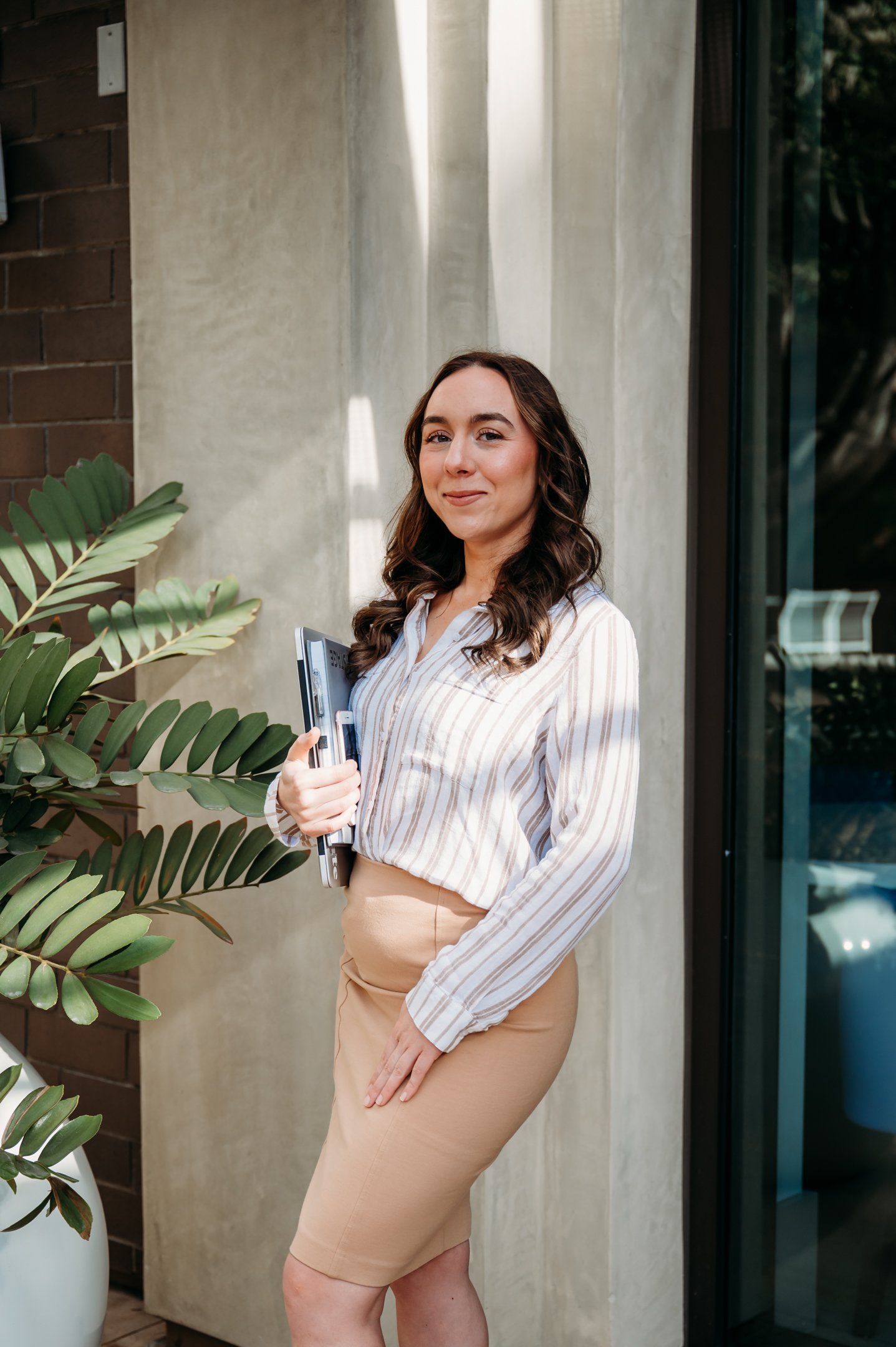 A woman with long curly brown hair, wearing a striped shirt and beige skirt, holding notebooks, standing outside near a concrete wall and a large green plant. Alisha Mowry Photography Military, Brand, and Portrait Photographer San Diego CA