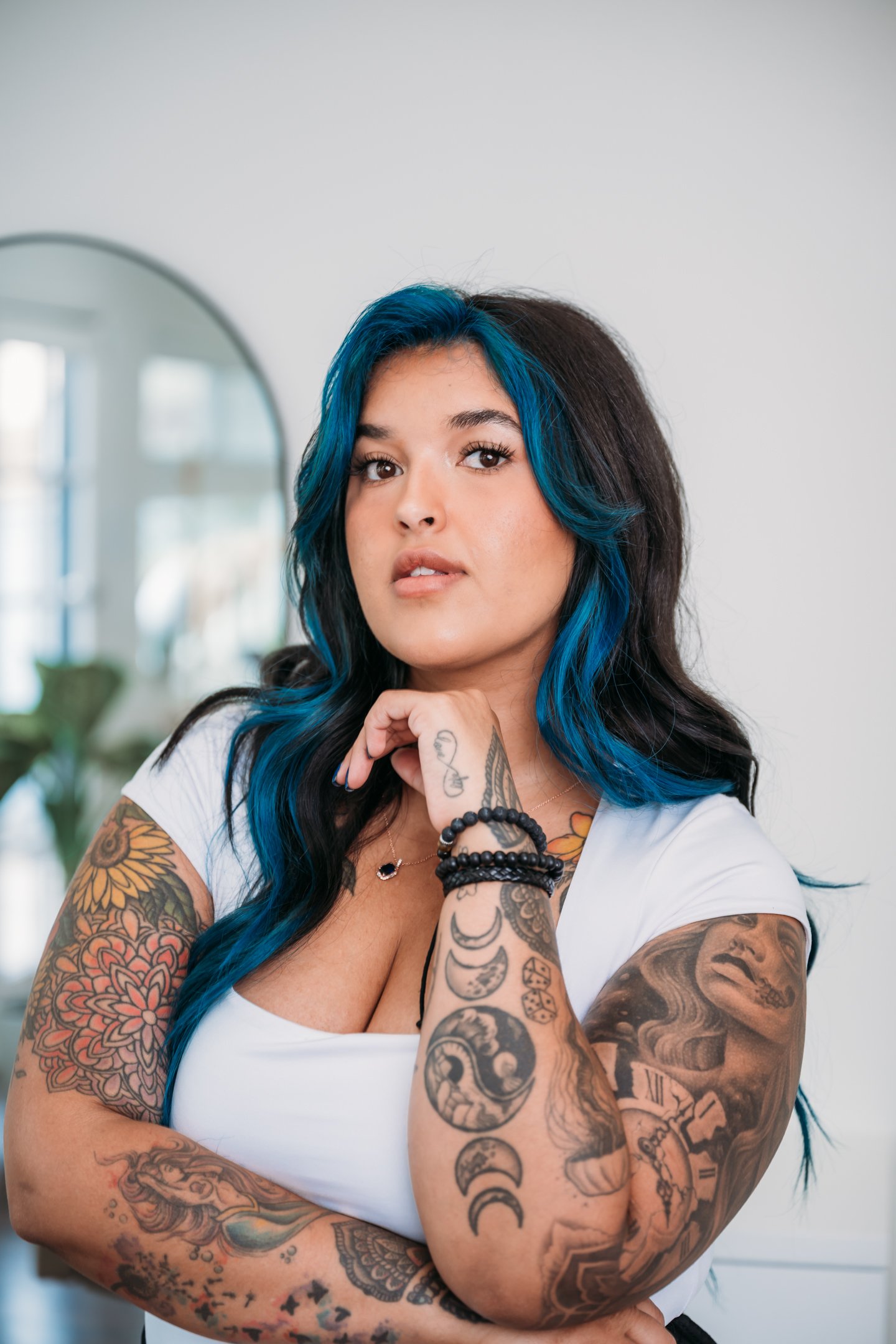 A woman with long, wavy black hair with blue streaks, wearing a white top, showing tattoos on her arms, chest, and hand, looking confidently at the camera. Alisha Mowry Photography Military, Brand, and Portrait Photographer San Diego CA