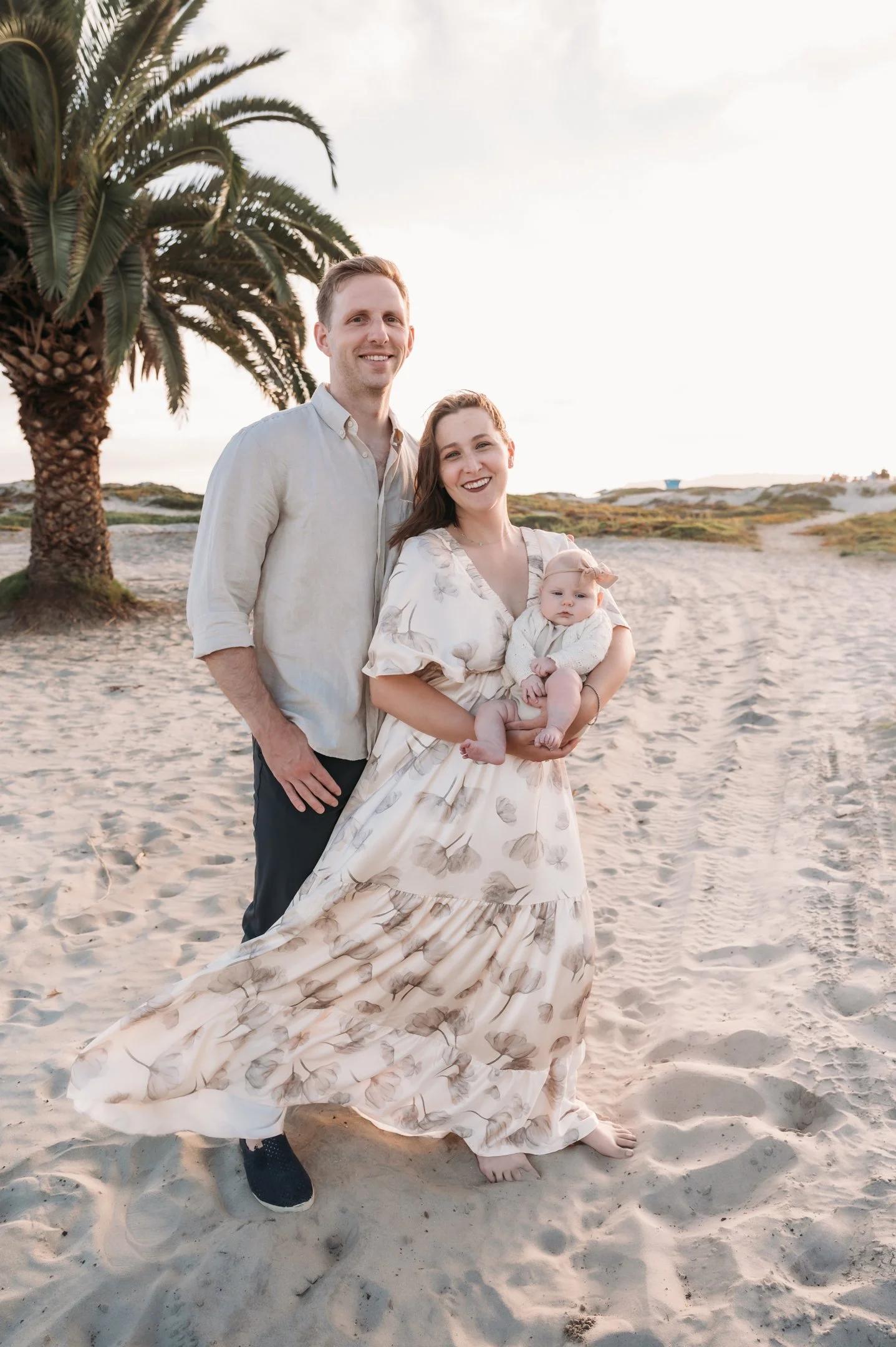 A family of three standing on a sandy beach near a palm tree, smiling at the camera during sunset.