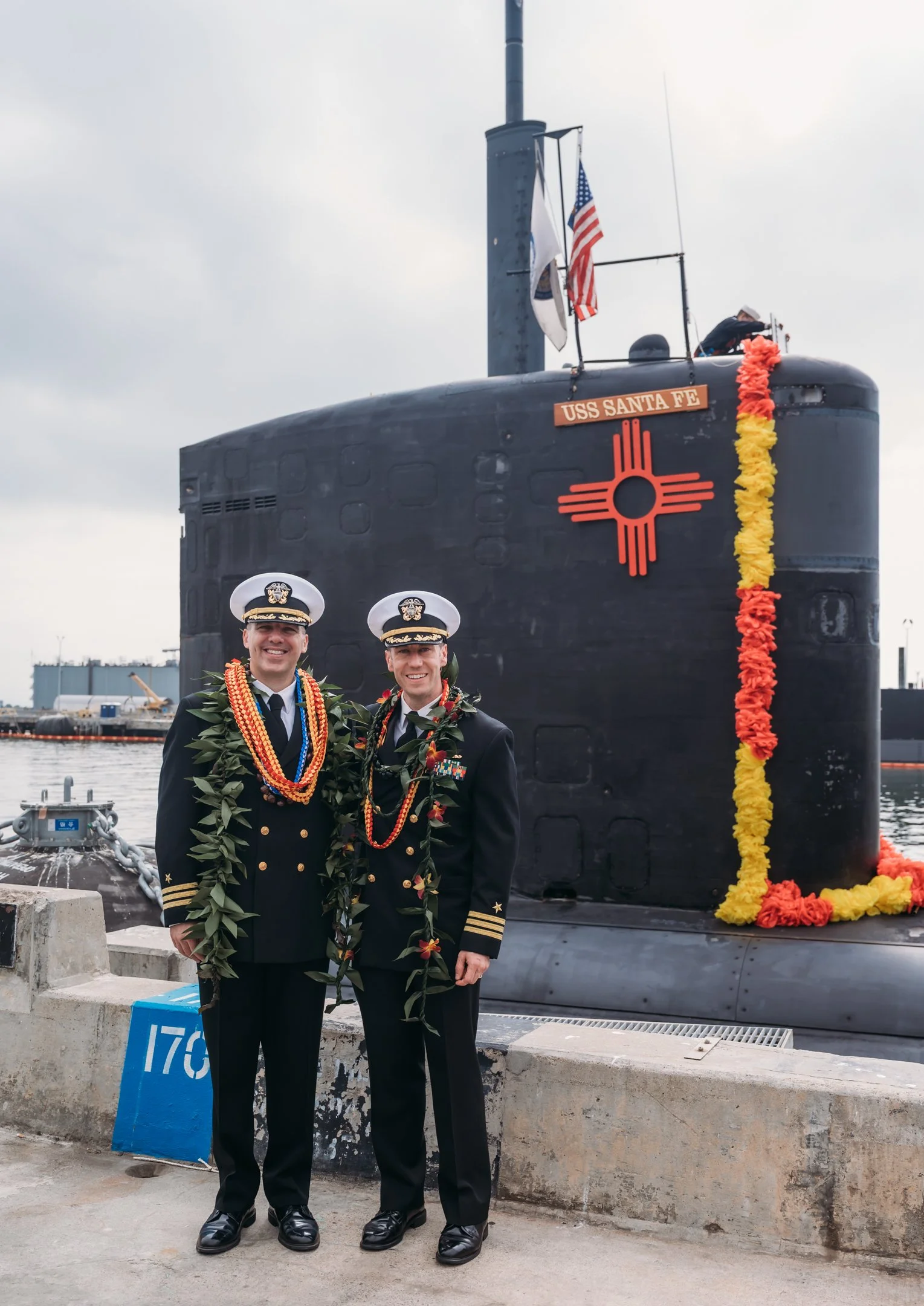 Two Navy officers in dress uniforms standing in front of a submarine named USS Santa Fe, decorated with flowers and national flags, during a ceremony. Alisha Mowry Photography Military Photographer San Diego