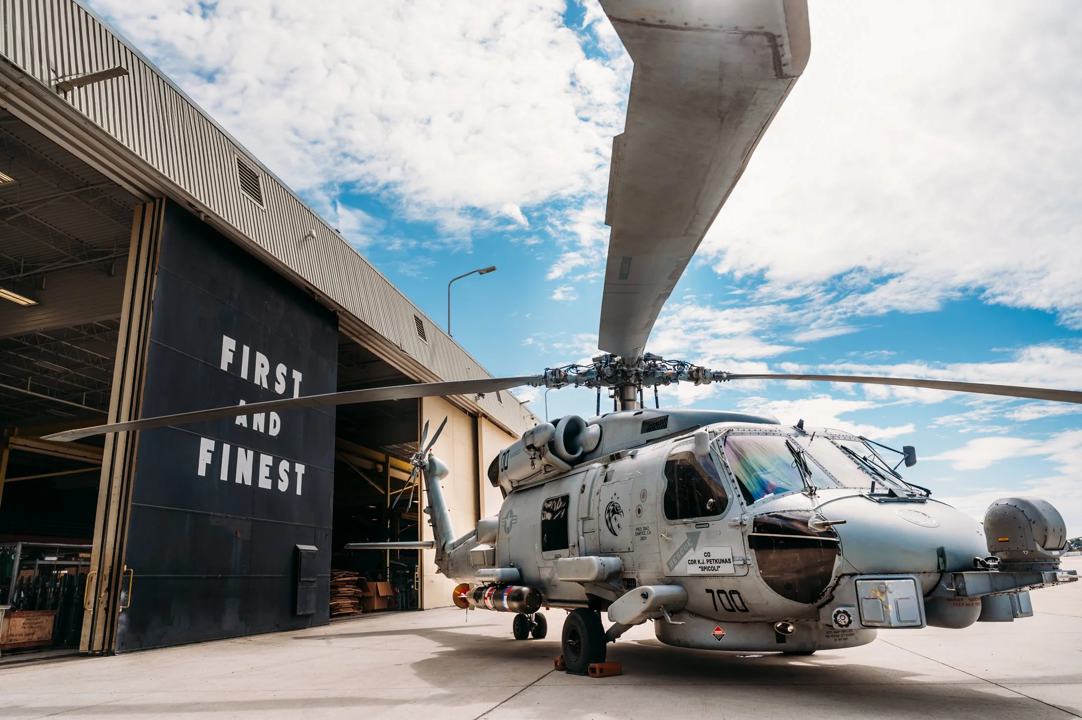 A military helicopter parked outside an aircraft hangar with the words 'First and Finest' on the hangar door, under a partly cloudy sky. Alisha Mowry Photography Military Photographer San Diego