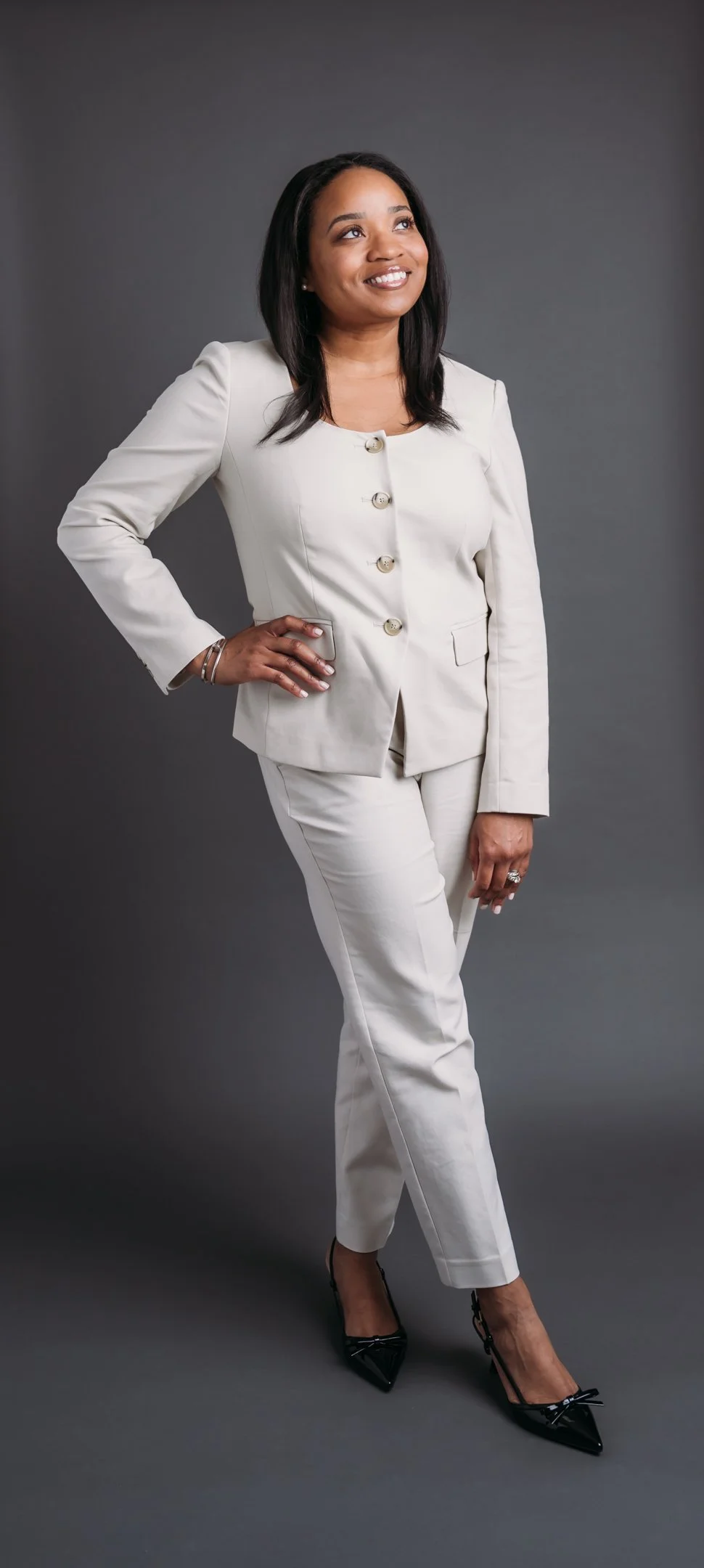 A woman in a white suit with buttons, standing against a dark gray background, smiling, with one hand on her hip and the other hanging down, wearing black pointed heels with bows. Alisha Mowry Photography Military, Brand, and Portrait Photographer Sa