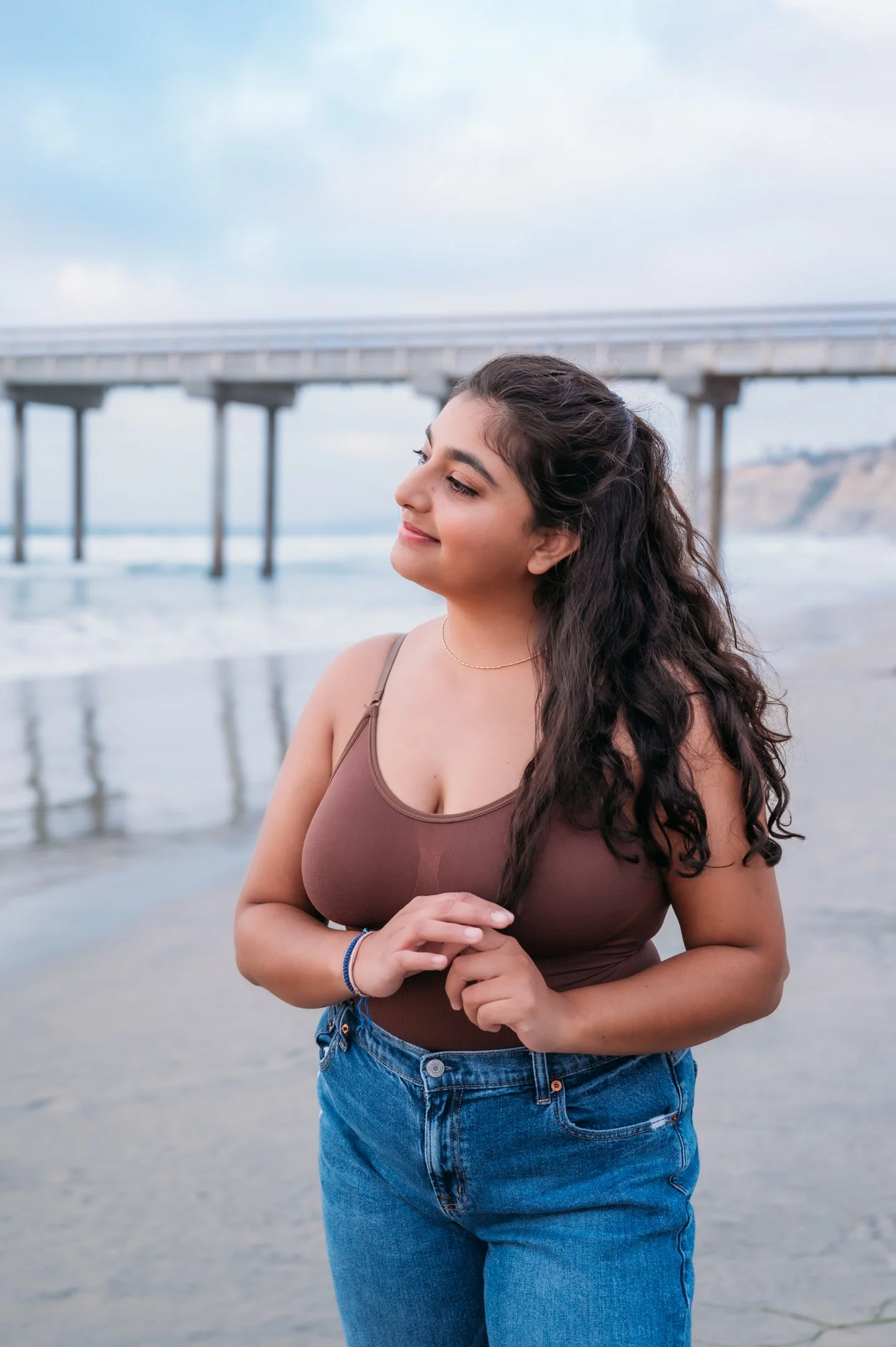 Young woman standing on a beach near the ocean, with a pier in the background, smiling and-looking away. Alisha Mowry Photography Military, Brand, and Portrait Photographer San Diego CA