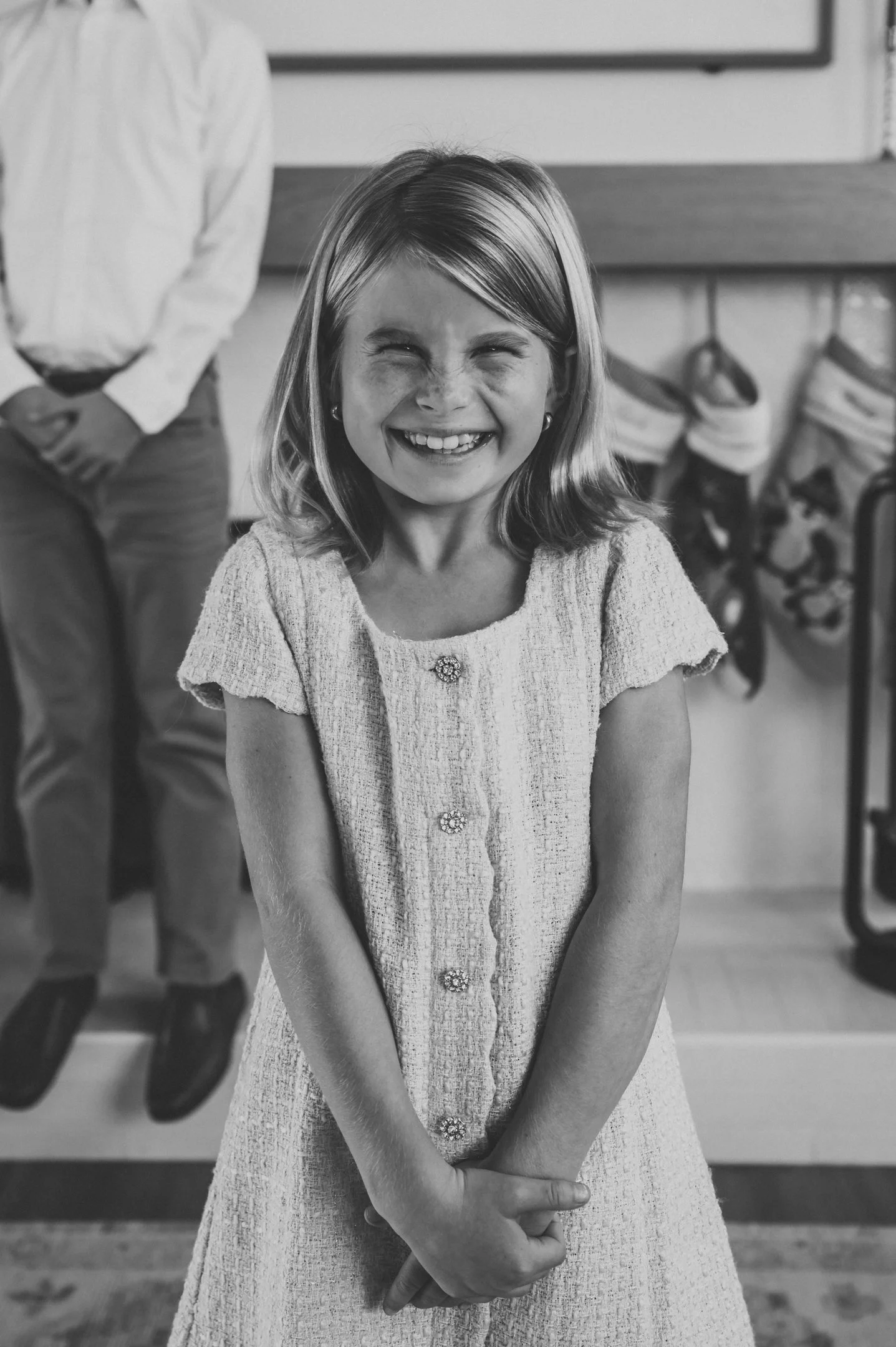 A young girl with freckles and shoulder-length hair smiling and clasping her hands in front of her, standing indoors with a Christmas stocking hanging on the wall in the background. Alisha Mowry Photography Military, Brand, and Portrait Photographer 