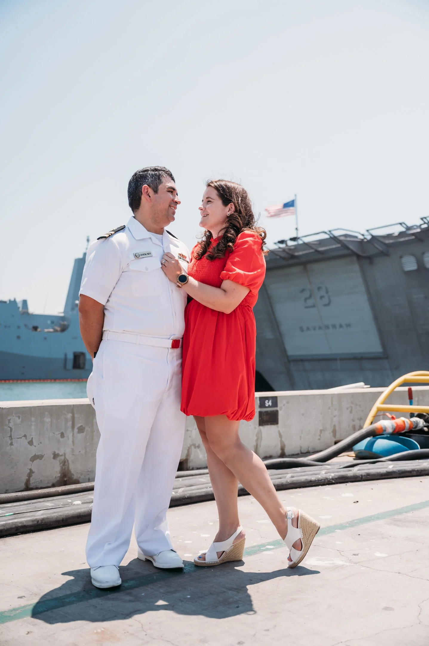 A couple, a man in a white military uniform and a woman in a red dress, standing and smiling at each other near a naval ship dock with an American flag. uss midway military retirement ceremony 