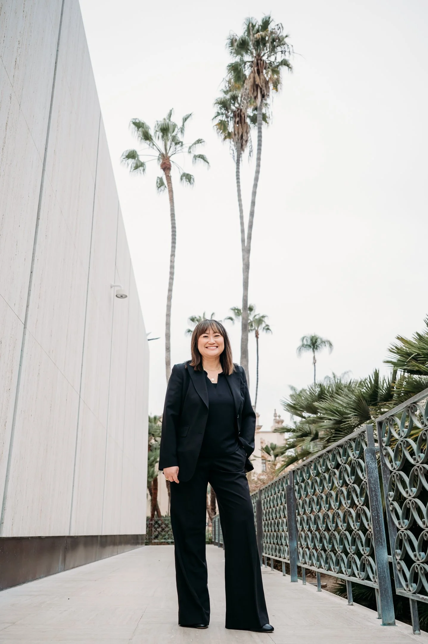 A woman dressed in a black suit smiling while standing outdoors on a sidewalk with palm trees in the background. Alisha Mowry Photography Military, Brand, and Portrait Photographer San Diego CA