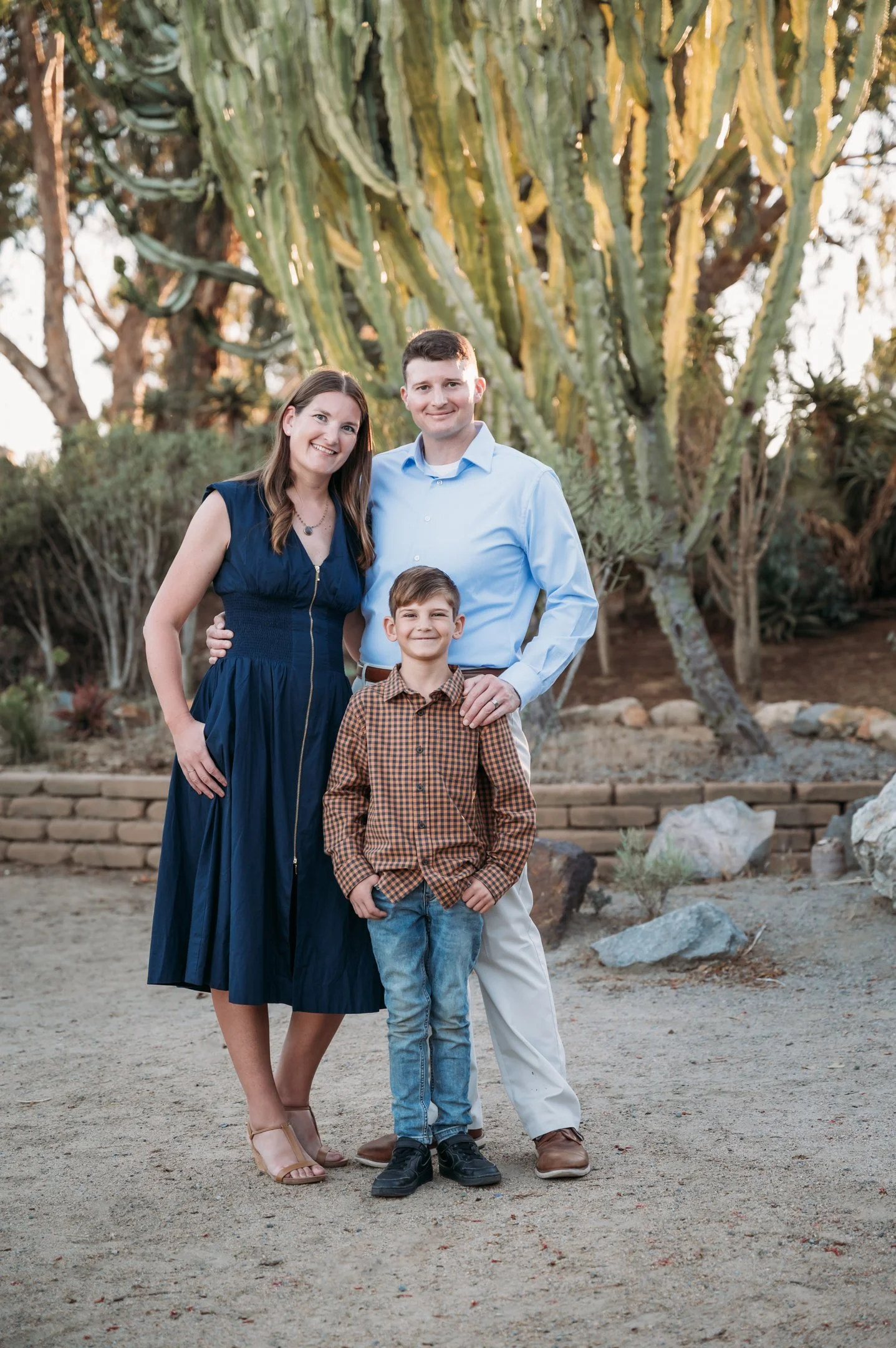 A family of three standing outdoors in front of a large cactus, posing for a photo. The mother is wearing a navy blue dress, the father is in a light blue shirt and white pants, and their young son is dressed in a brown checkered shirt and jeans. The