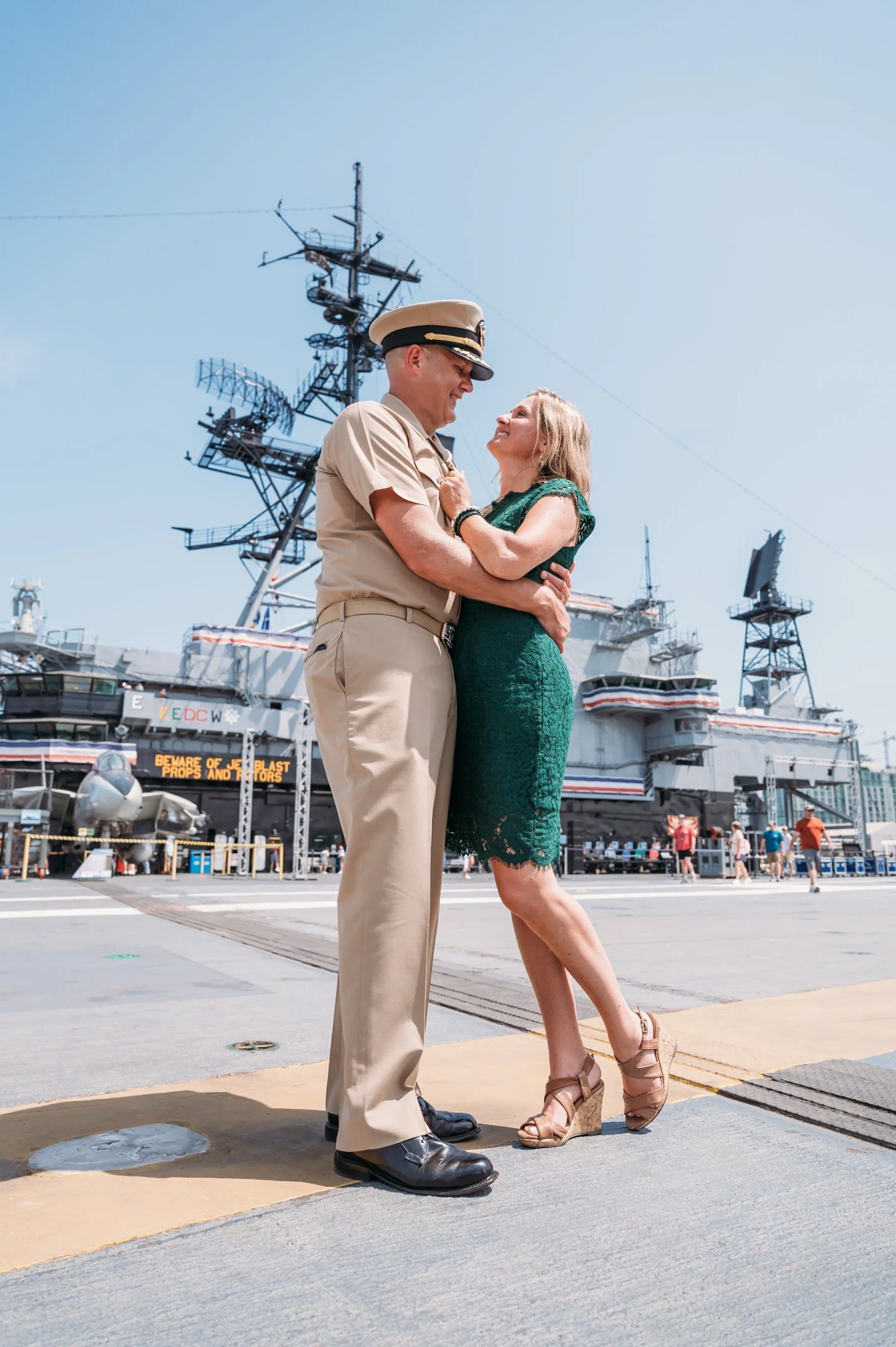 A couple dancing outdoors near a large military ship, with the man dressed in a Navy uniform and the woman in a green dress. uss midway military retirement ceremony 
