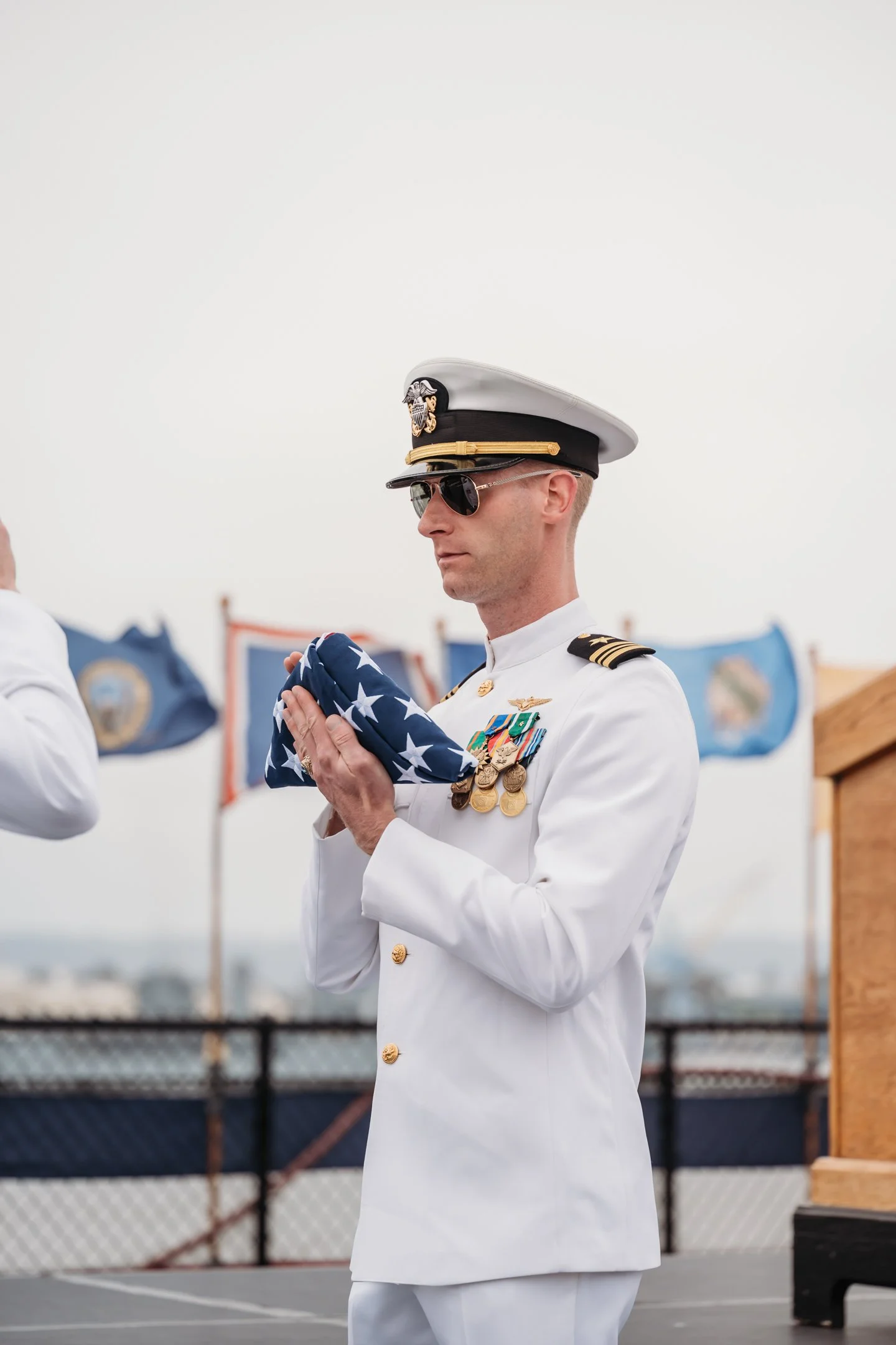 A man in a white military dress uniform holding a folded American flag during a ceremony, with patriotic flags in the background. USS Midway retirement ceremony by Alisha Mowry Photography 