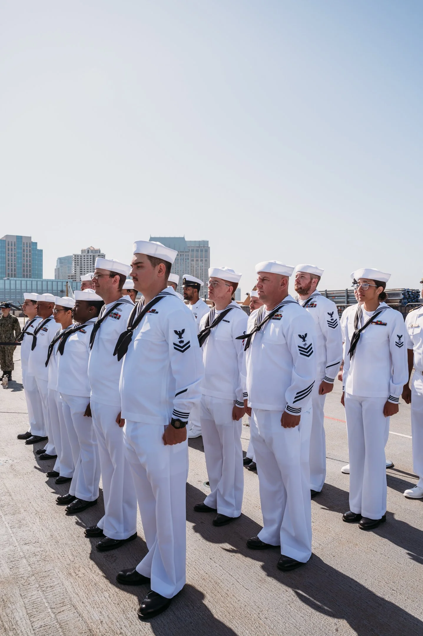 A group of U.S. Navy sailors standing in formation outdoors, wearing white uniforms and hats, with a city skyline in the background. Alisha Mowry Photography Military Photographer San Diego