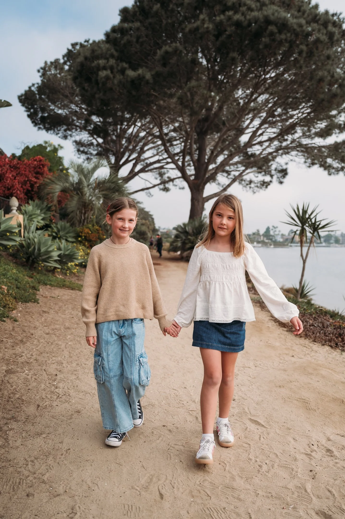 Two girls walking hand-in-hand on a dirt path near water, with trees and plants in the background. Alisha Mowry Photography Military, Brand, and Portrait Photographer San Diego CA