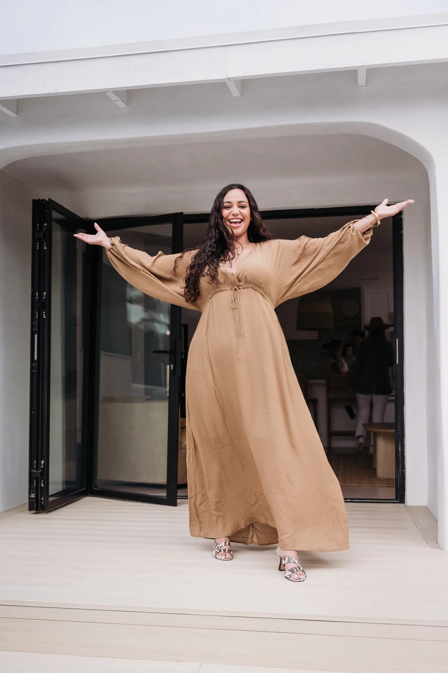 Woman in tan dress standing on a porch with arms outstretched, smiling. Alisha Mowry Photography Military, Brand, and Portrait Photographer San Diego CA