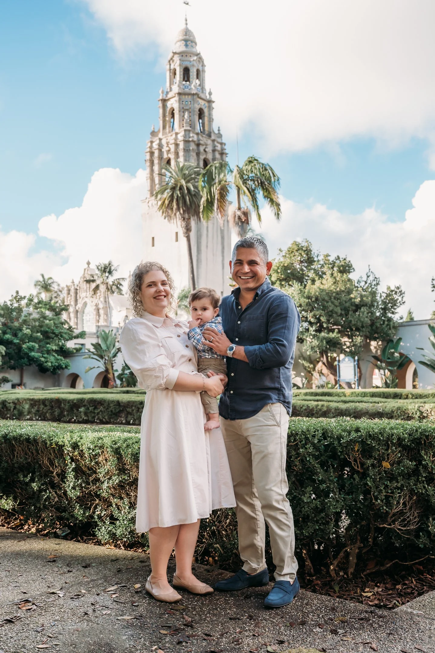 A family of three standing outdoors in front of a historic building with a tall, ornate tower, palm trees, and lush greenery. The woman is holding a young child, and a man stands beside them, all smiling. Alisha Mowry Photography Military, Brand, and