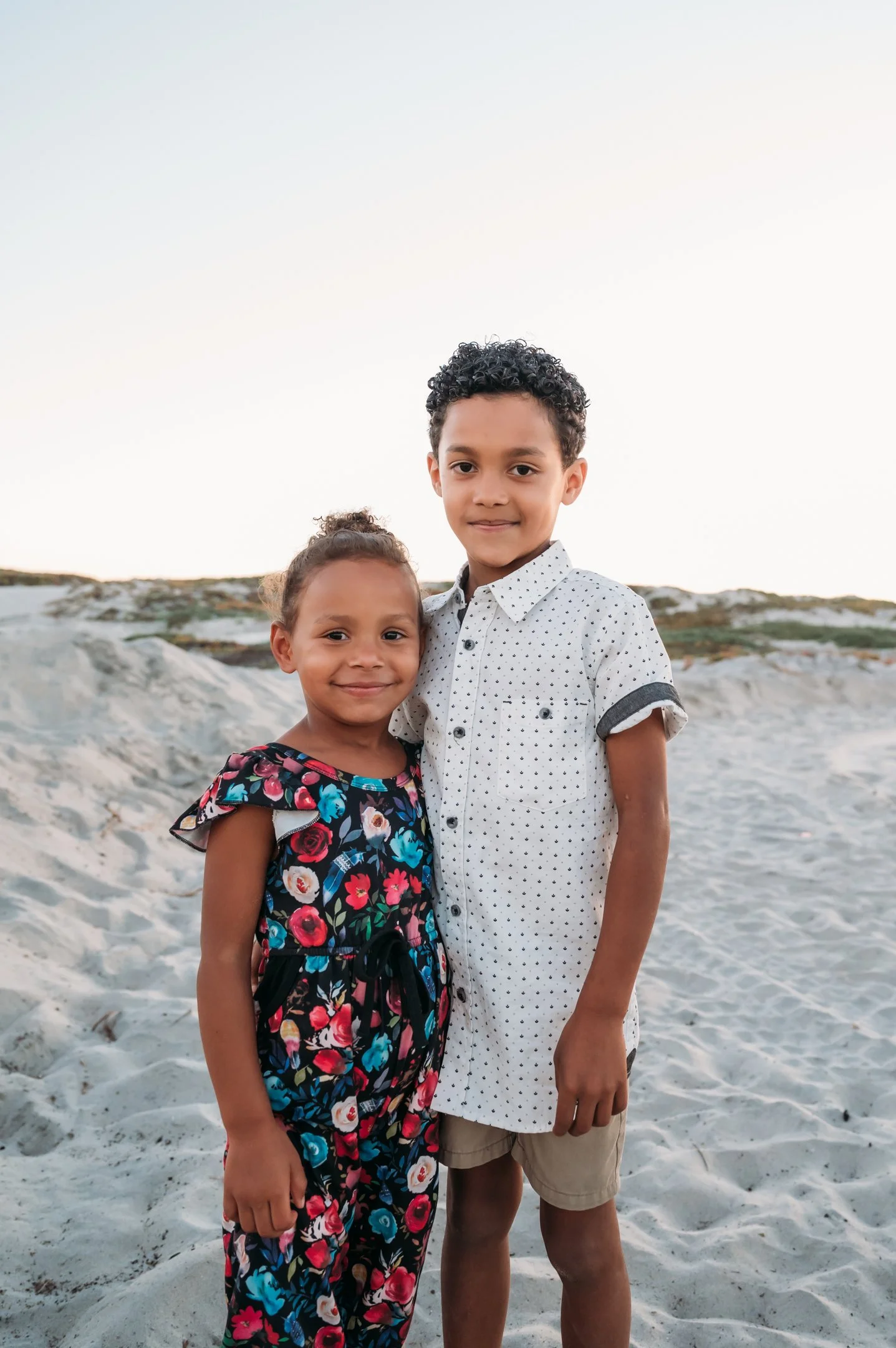 A young boy and girl standing together on a sandy beach, smiling at the camera during sunset. Alisha Mowry Photography Military, Brand, and Portrait Photographer San Diego CA