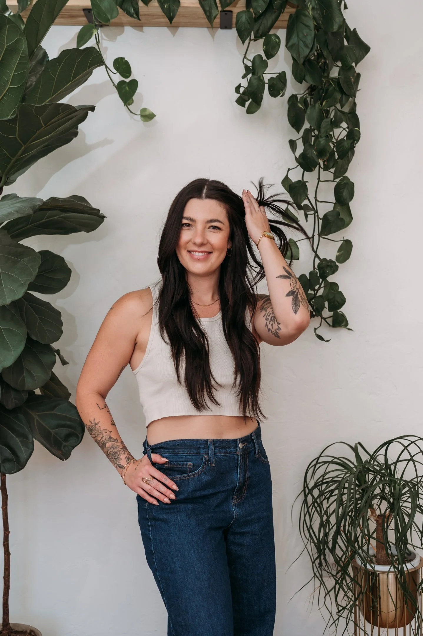 A woman with long dark hair smiling, wearing a white crop top and jeans, standing among large indoor plants against a white wall. Alisha Mowry Photography Military, Brand, and Portrait Photographer San Diego CA