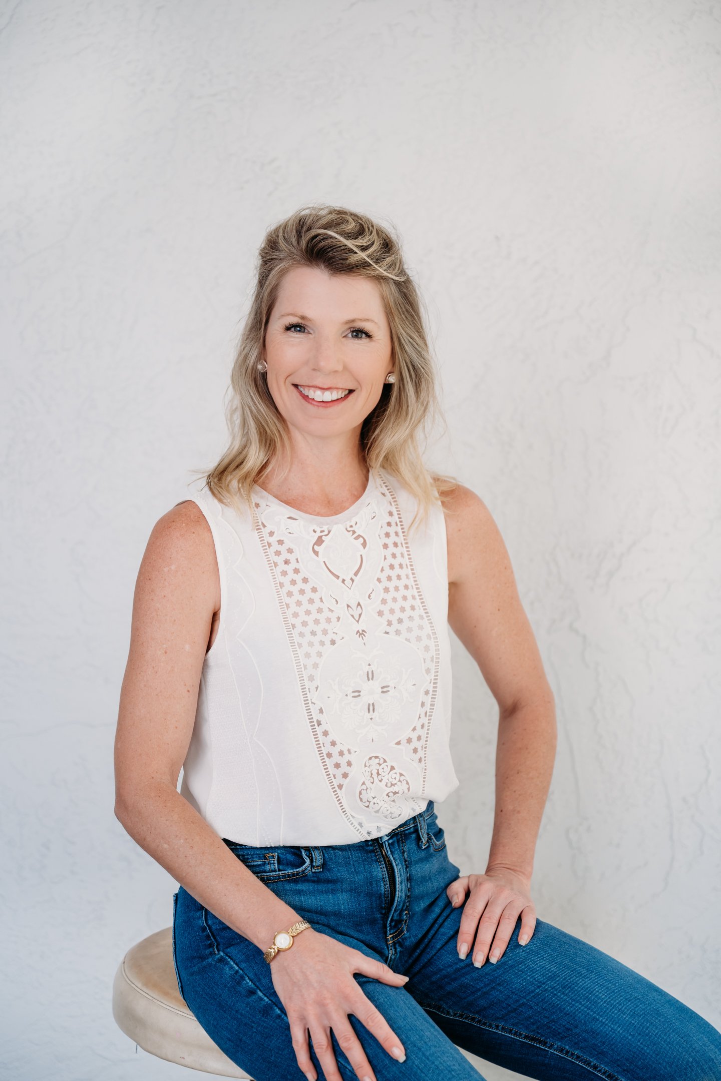 Smiling woman with blonde hair wearing a white sleeveless embroidered top and blue jeans, sitting on a beige stool against a white textured background. Alisha Mowry Photography Military, Brand, and Portrait Photographer San Diego CA