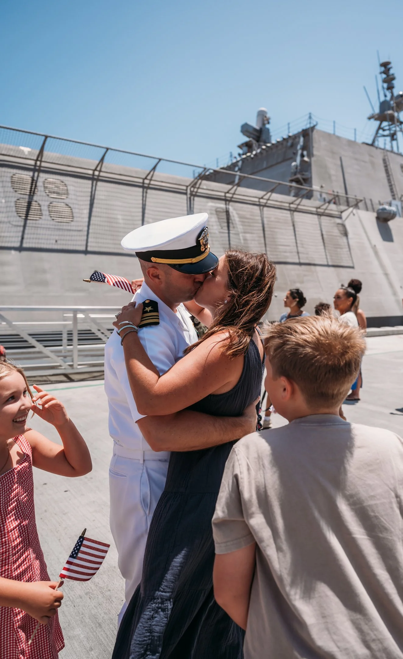 A couple, the woman in a dark dress and the man in a white navy uniform, are kissing on an aircraft carrier with children and other people in the background. The girl is holding a small American flag. Alisha Mowry Photography Military Photographer Sa