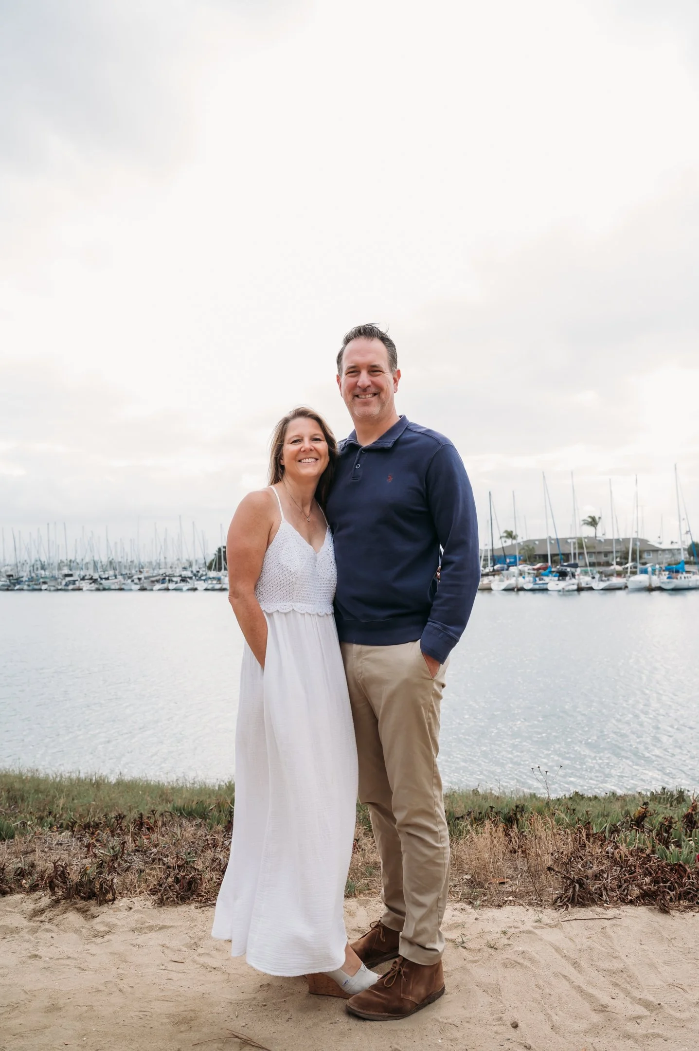 A smiling couple standing on a sandy bank by a marina with sailboats, overcast sky, and distant trees. Alisha Mowry Photography Military, Brand, and Portrait Photographer San Diego CA