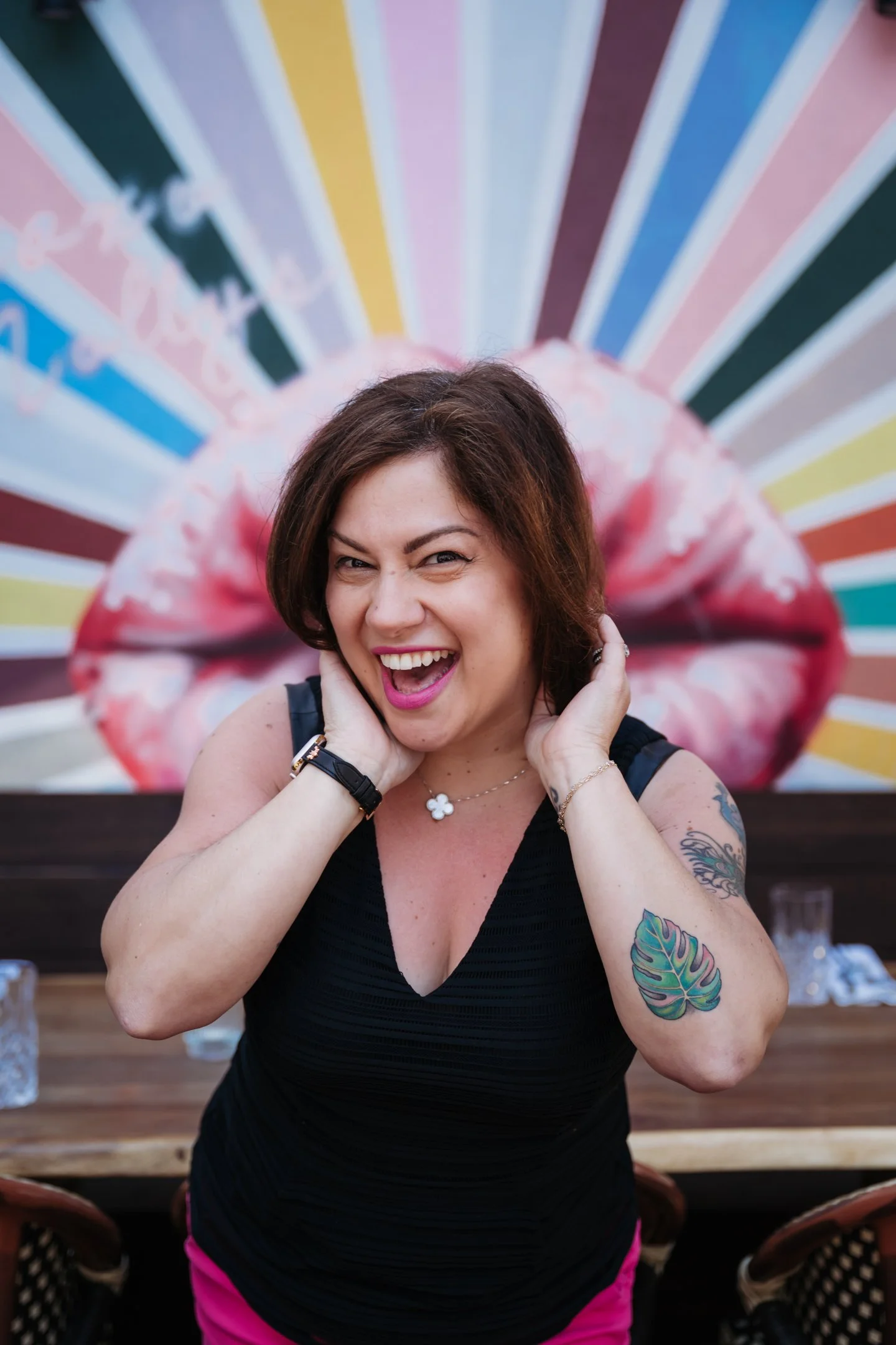 A woman with dark brown hair, wearing a black sleeveless top and pink pants, is smiling and playfully winking in front of a colorful mural with large lips and radiating stripes. Alisha Mowry Photography Military, Brand, and Portrait Photographer San 