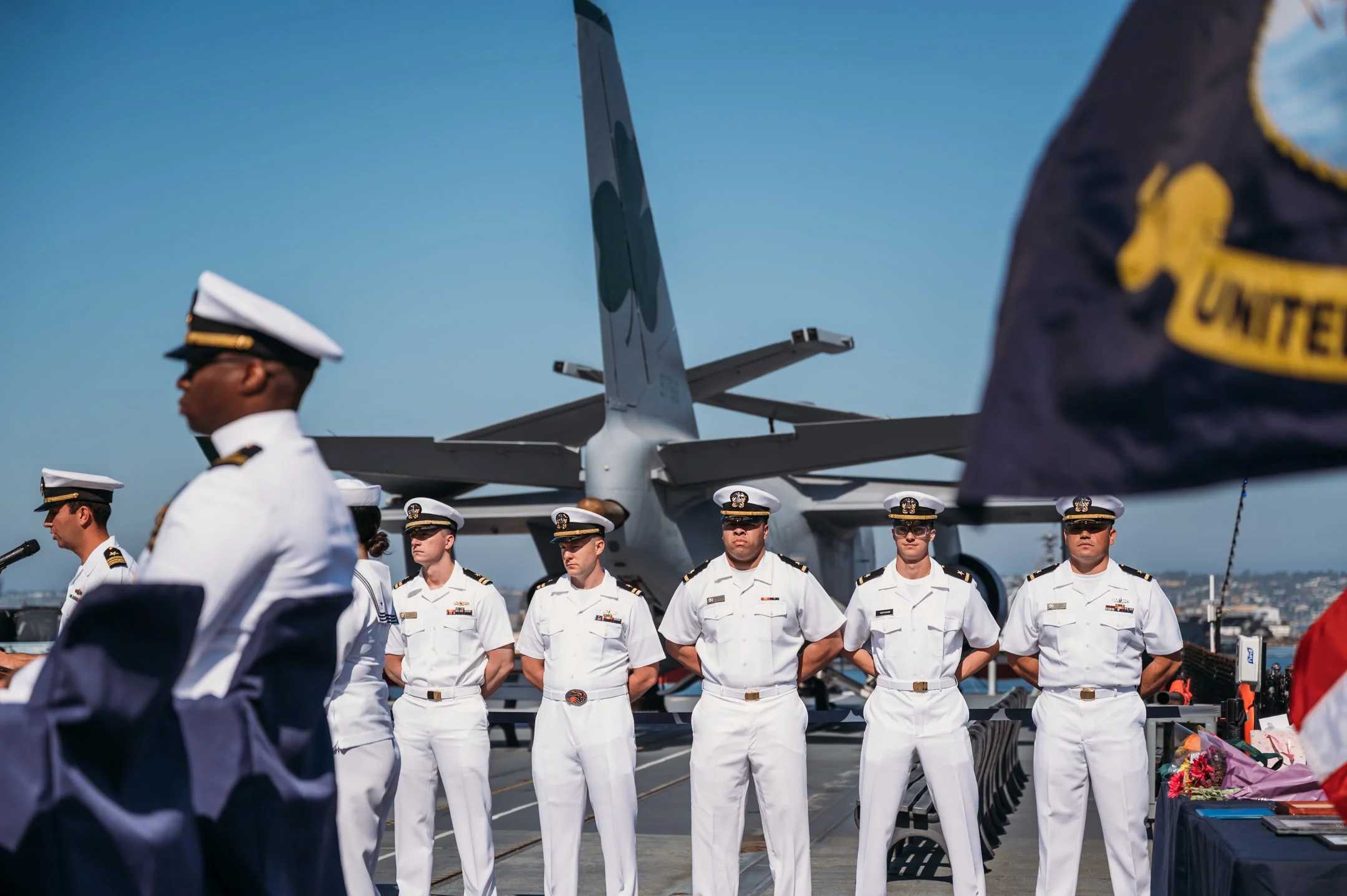 U.S. Navy officers in white uniforms standing in formation on an aircraft carrier deck, with a fighter jet in the background and a blue sky overhead.  Alisha Mowry Photography Military Photographer San Diego